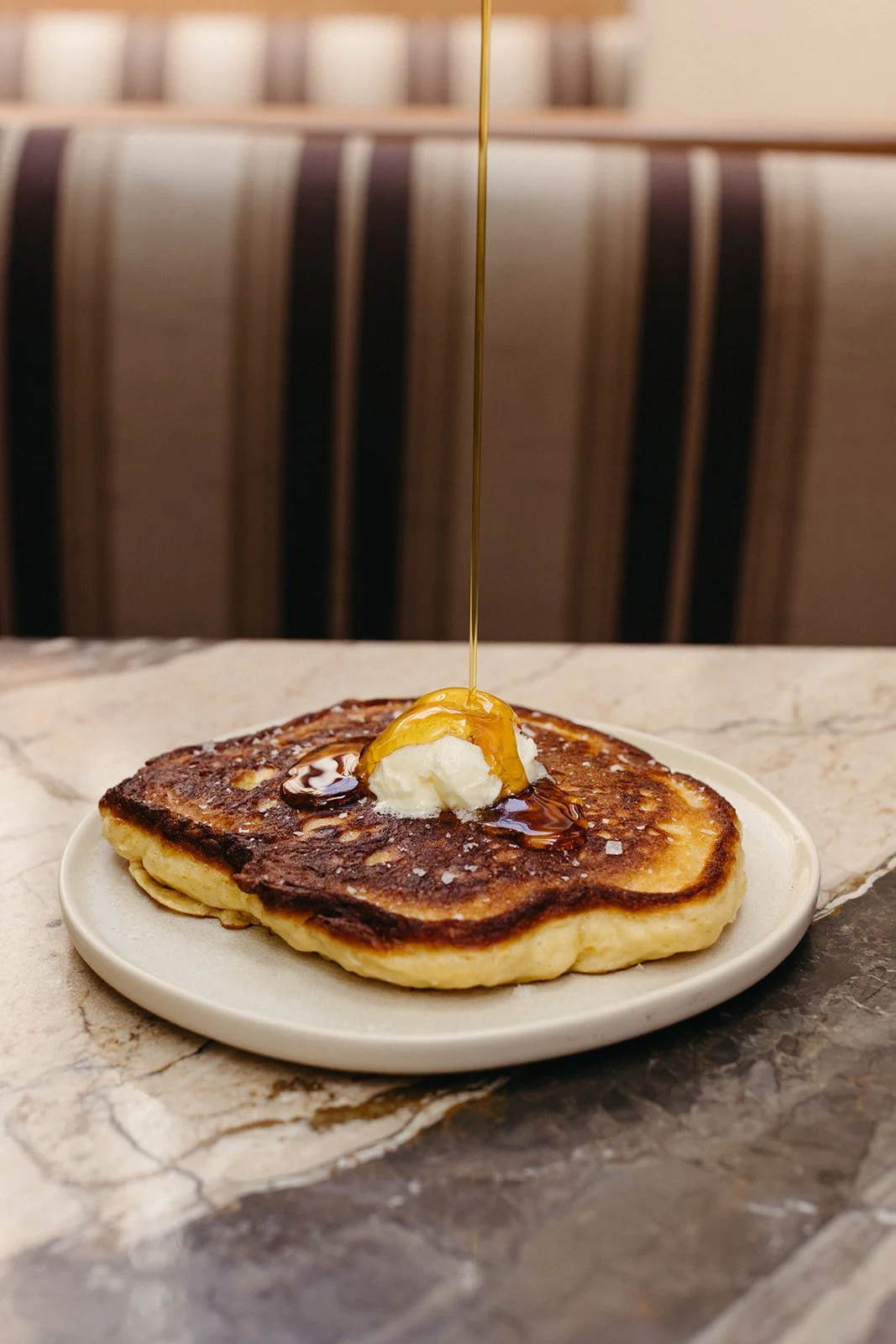 Plate with a golden-brown pancake topped with butter, syrup, and a drizzle of honey, on a marble table with a blurred background of striped chairs.