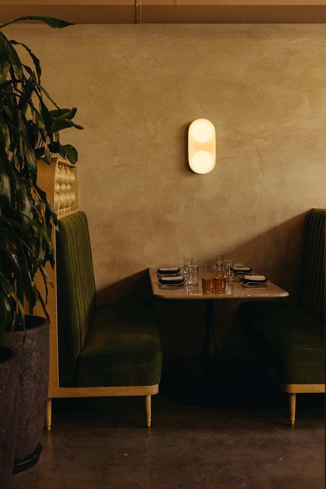 A cozy restaurant booth with green upholstered seats and a marble table, set with glasses and napkins. A wall-mounted light fixture is above the table, and a large plant is on the left side of the image.