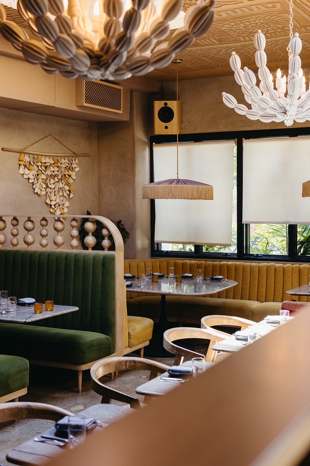 Interior of a cozy restaurant with green and mustard chairs, wooden tables set with glasses and napkins, decorative light fixtures, and large windows with white blinds.