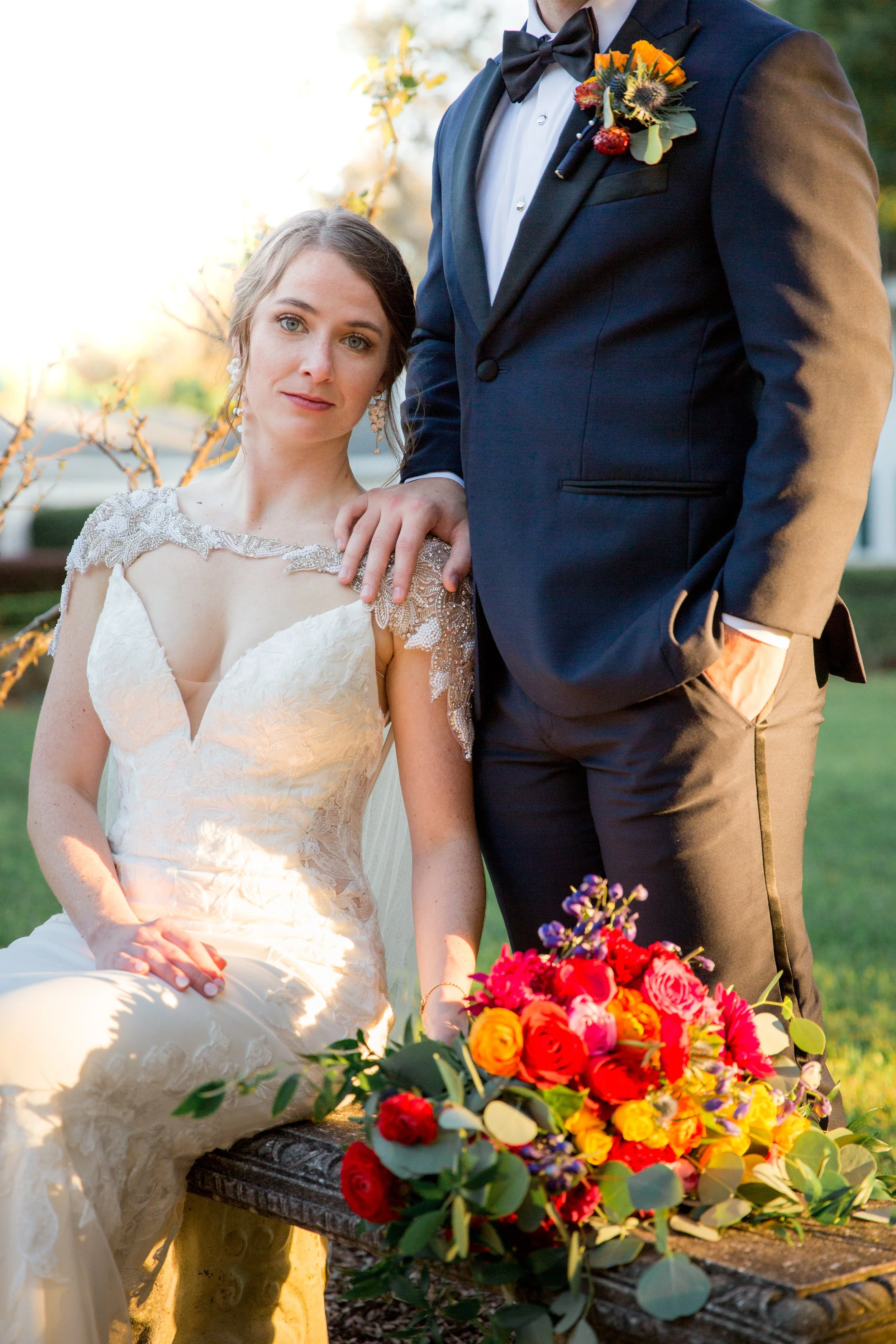 A bride and groom at their wedding, with the bride wearing a lace wedding gown and the groom in a tuxedo. They are outdoors with a colorful bouquet of flowers in front of them. The bride is sitting, and the groom is standing beside her, resting his hand on her shoulder.