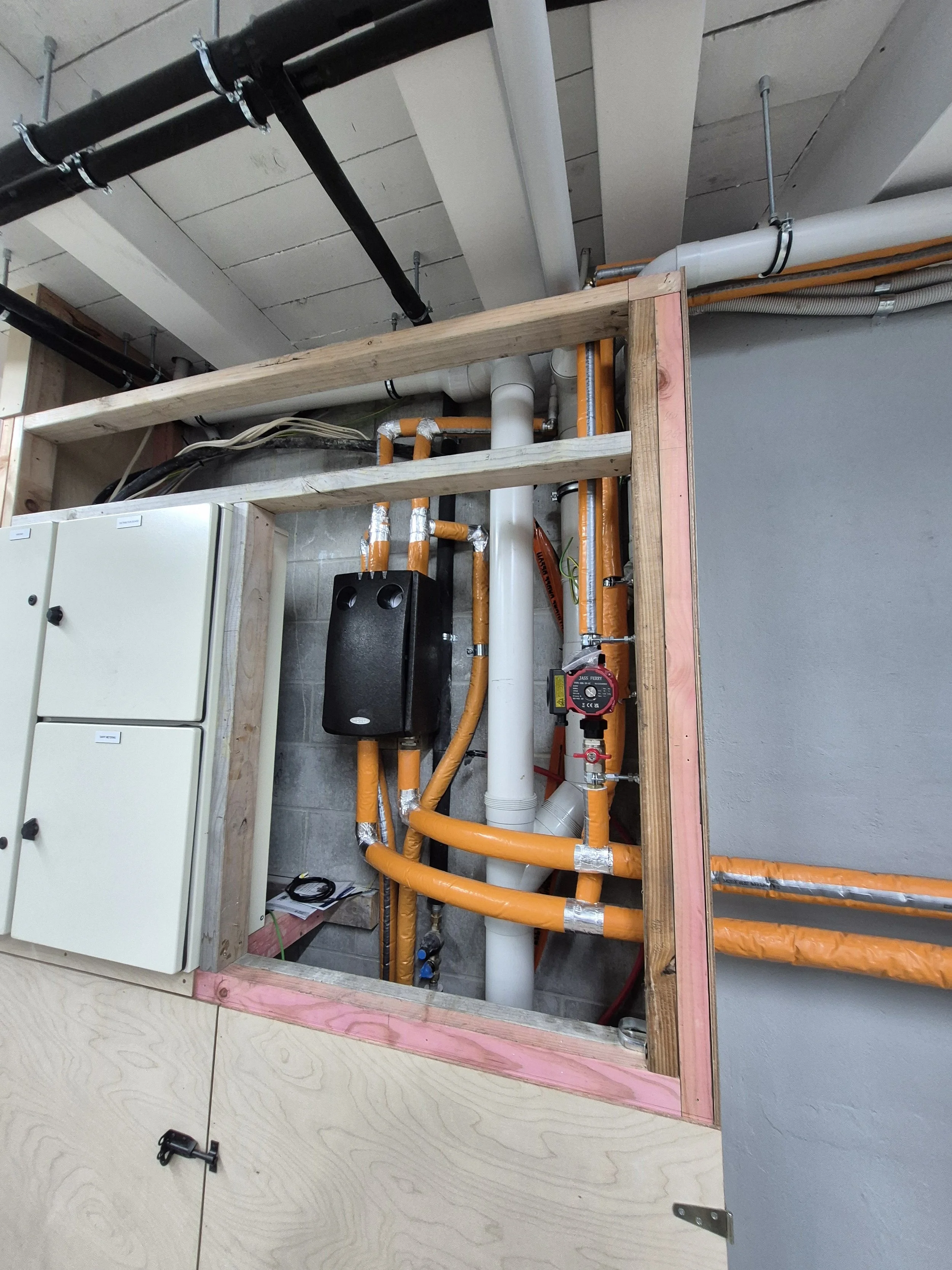 Construction area with plumbing pipes, orange insulation, a white vertical pipe, black speaker, electrical wires, and a wooden frame.