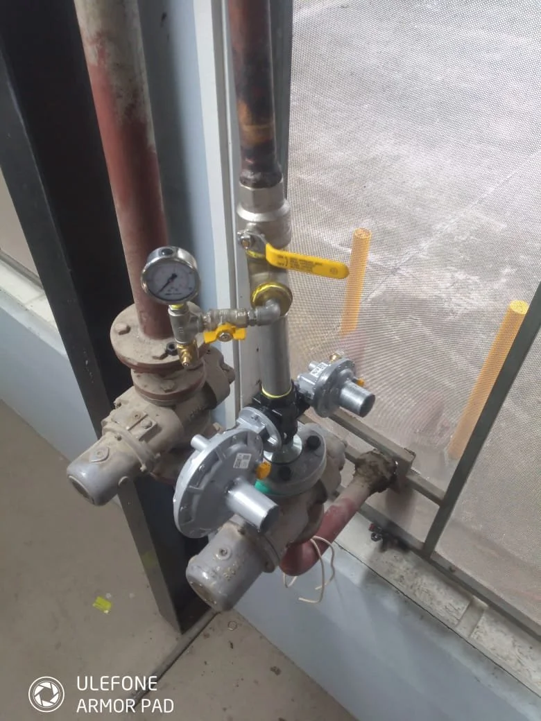 Industrial gas setup with gauges, valves, and pipes near a screened window.