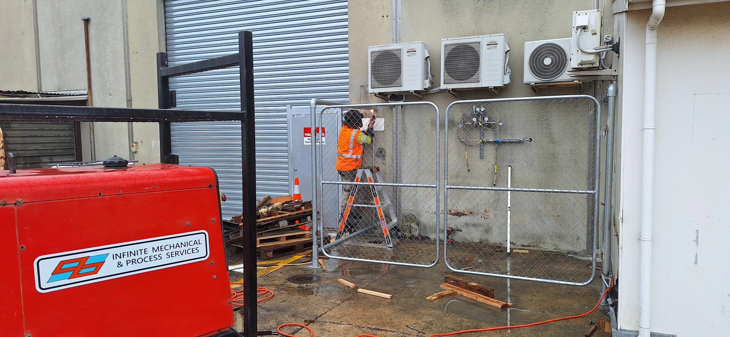A worker wearing an orange safety vest and yellow shirt is standing on a ladder, working on an electrical box or panel on a beige wall outside a building. There are three air conditioning units mounted on the wall above. A security fence surrounds th