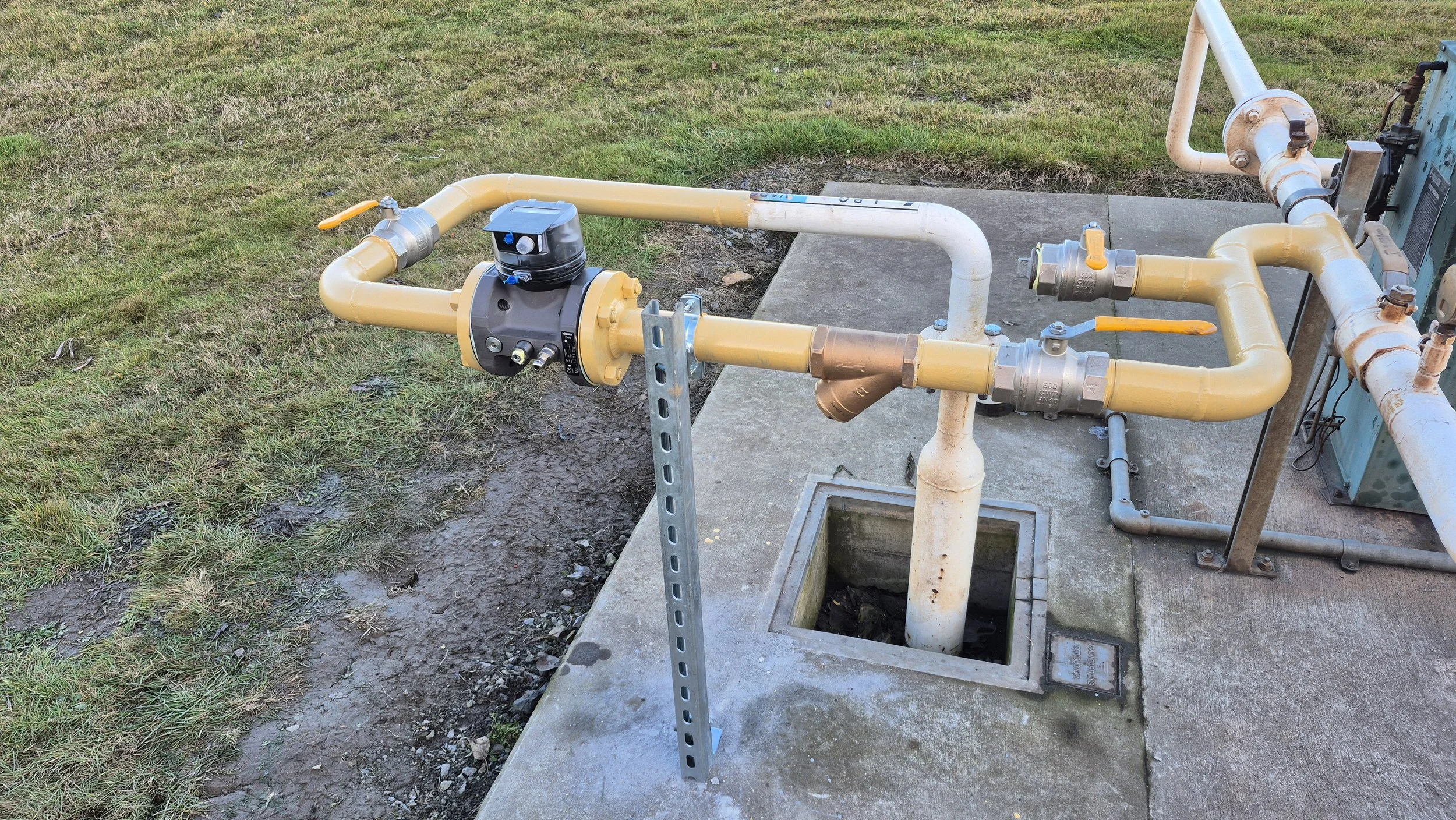 An outdoor water pipe system with yellow pipes, valves, and meters on a concrete pad next to a grassy area.
