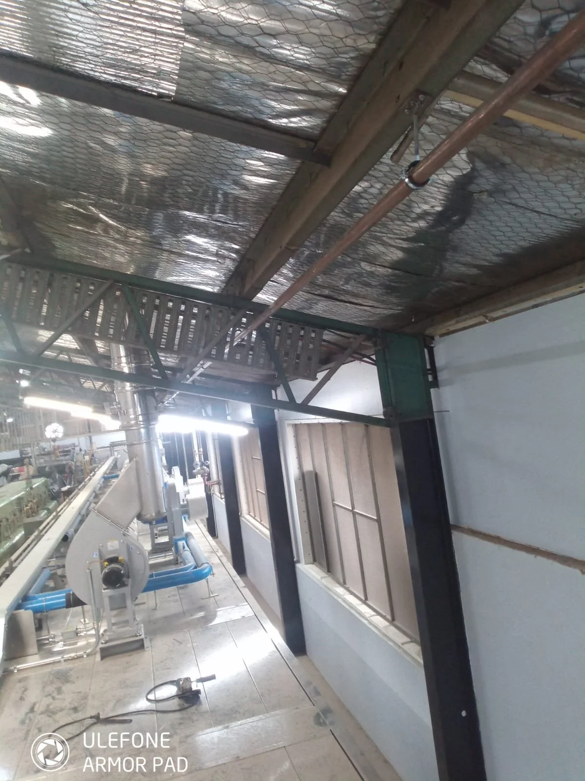 Industrial or factory setting with metal pipes, ducts, and machinery, and a window on the wall.
