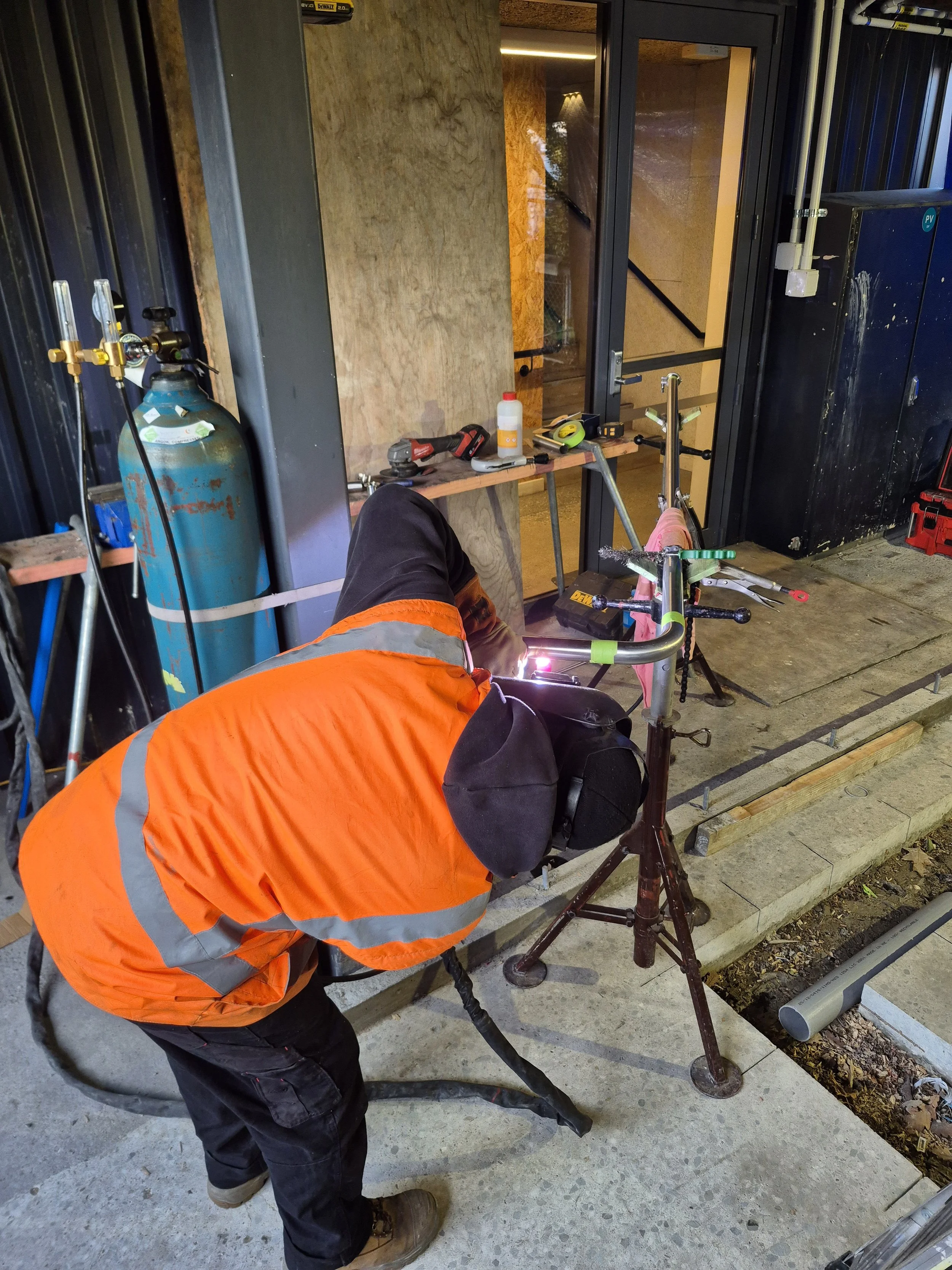 A construction worker wearing an orange safety jacket and brown boots is welding metal pipes outside a building. The worker is bent over, working on the pipes, which are positioned on the ground. A workbench with tools and a container is nearby.