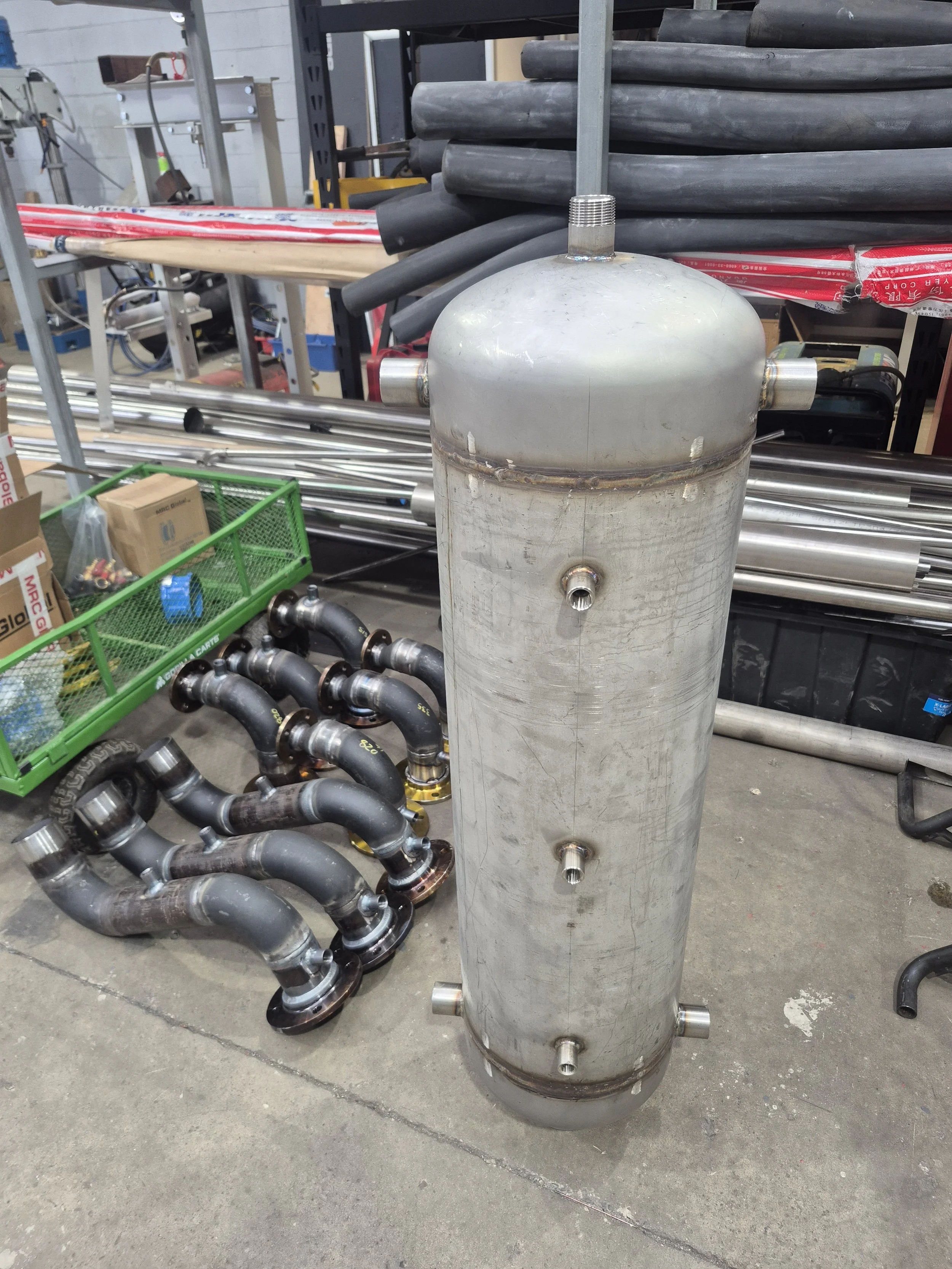 Metal cylinder with various pipes and equipment around it in a workshop or industrial setting.