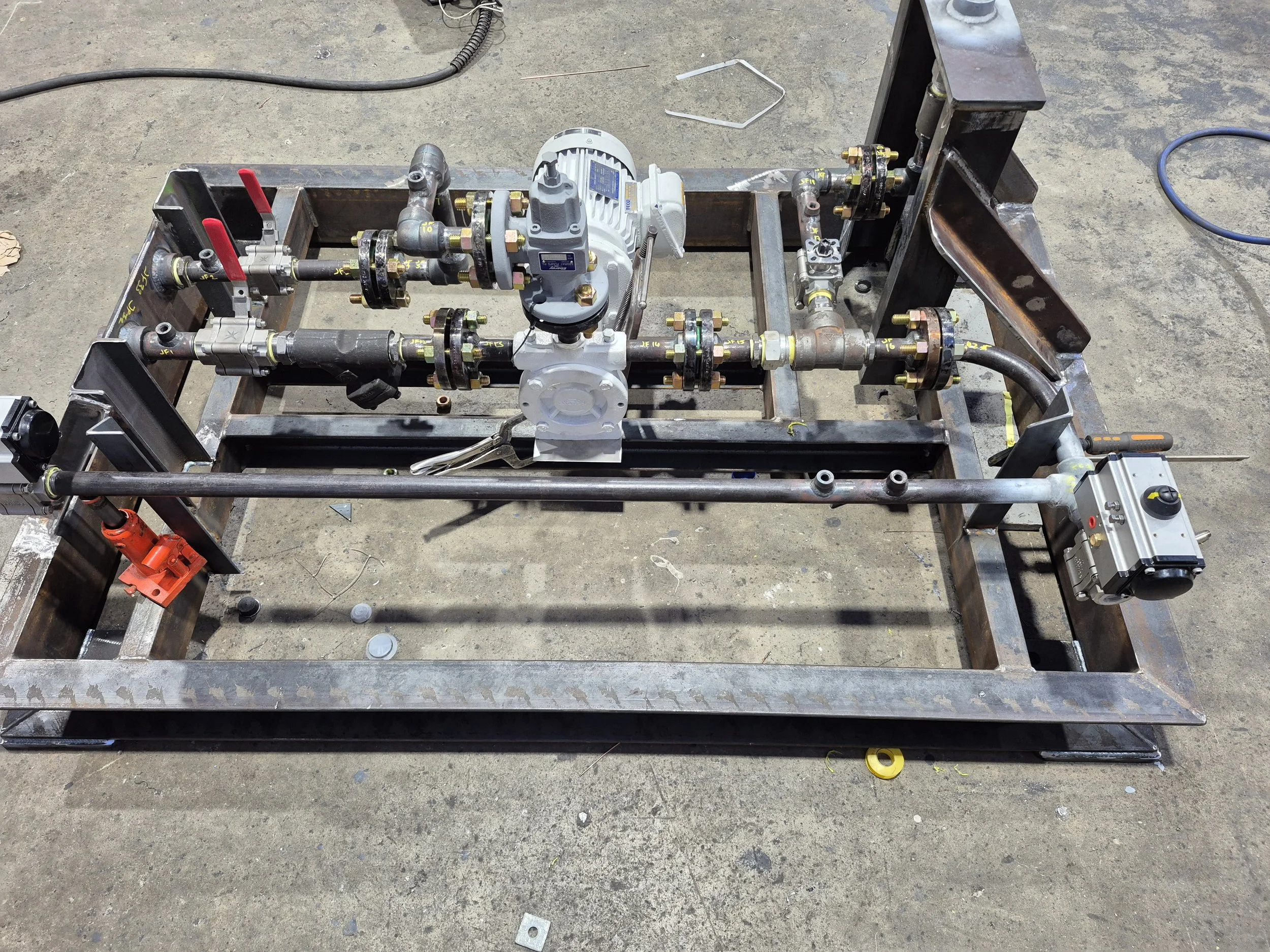 An industrial pipeline system mounted on a metal frame, with various pipes, valves, and a pump, situated on a concrete floor.