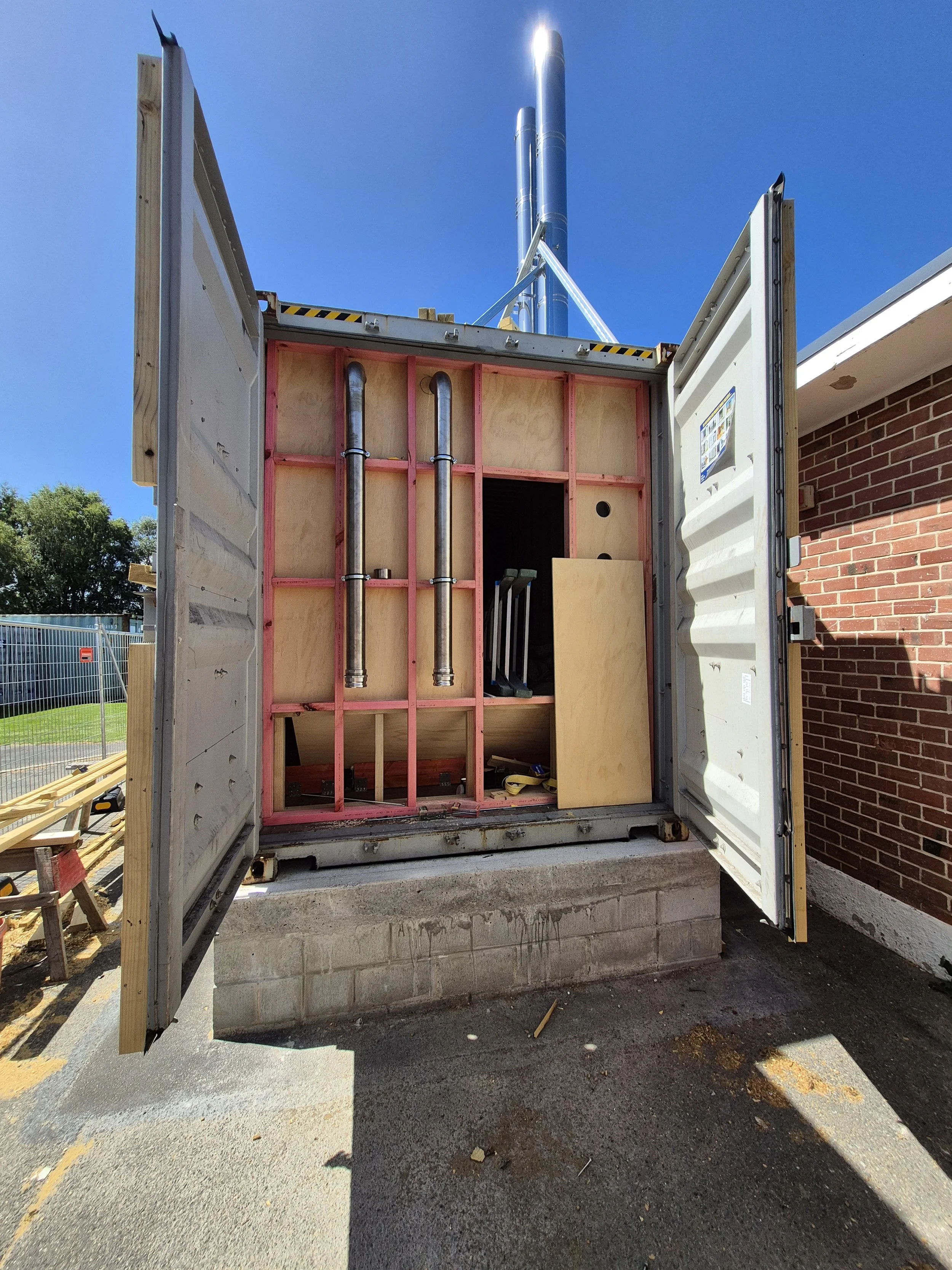 Open electrical or utility box with internal piping and wiring, mounted on a concrete block, positioned next to a brick building on a sunny day.