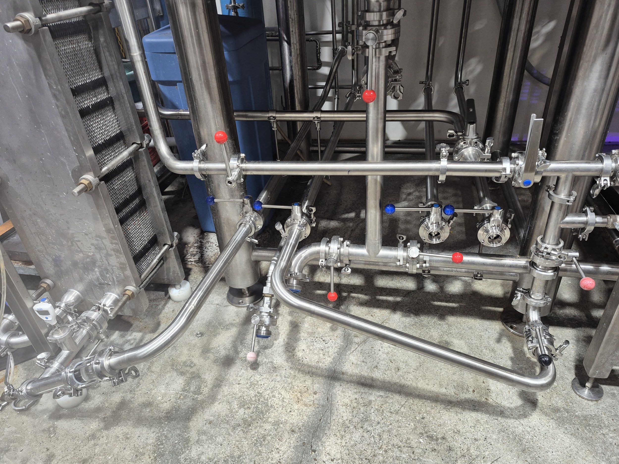 Stainless steel piping system with valves and red and blue knobs, mounted on a concrete floor, part of industrial machinery.