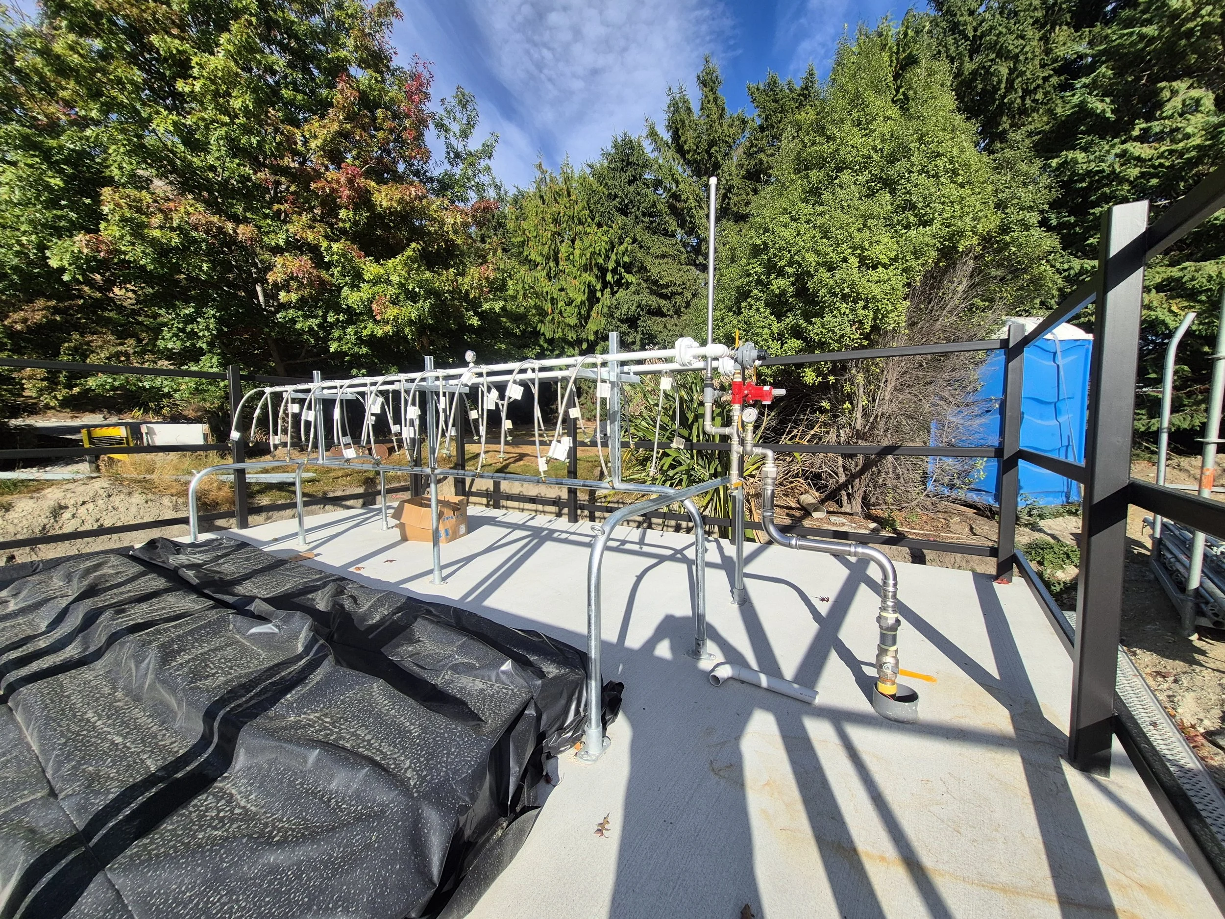 Construction site with piping, metal railing, blue portable toilet, and black plastic covering on a concrete surface, surrounded by trees and blue sky.