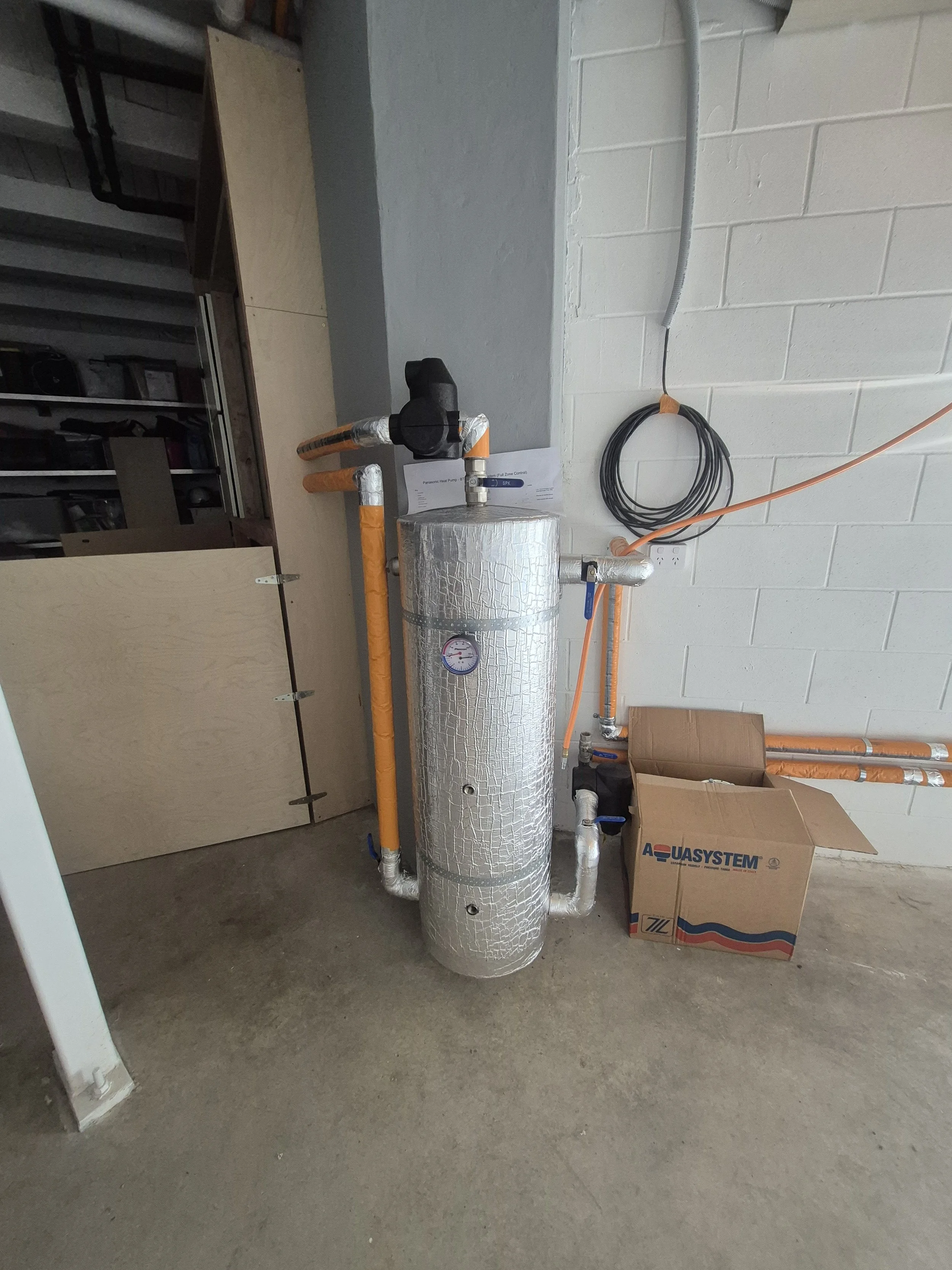 A large cylindrical heater wrapped in foil insulation, connected to orange and gray piping, situated against a white cinder block wall. There are cardboard boxes nearby and a coil of black cable hanging on the wall.