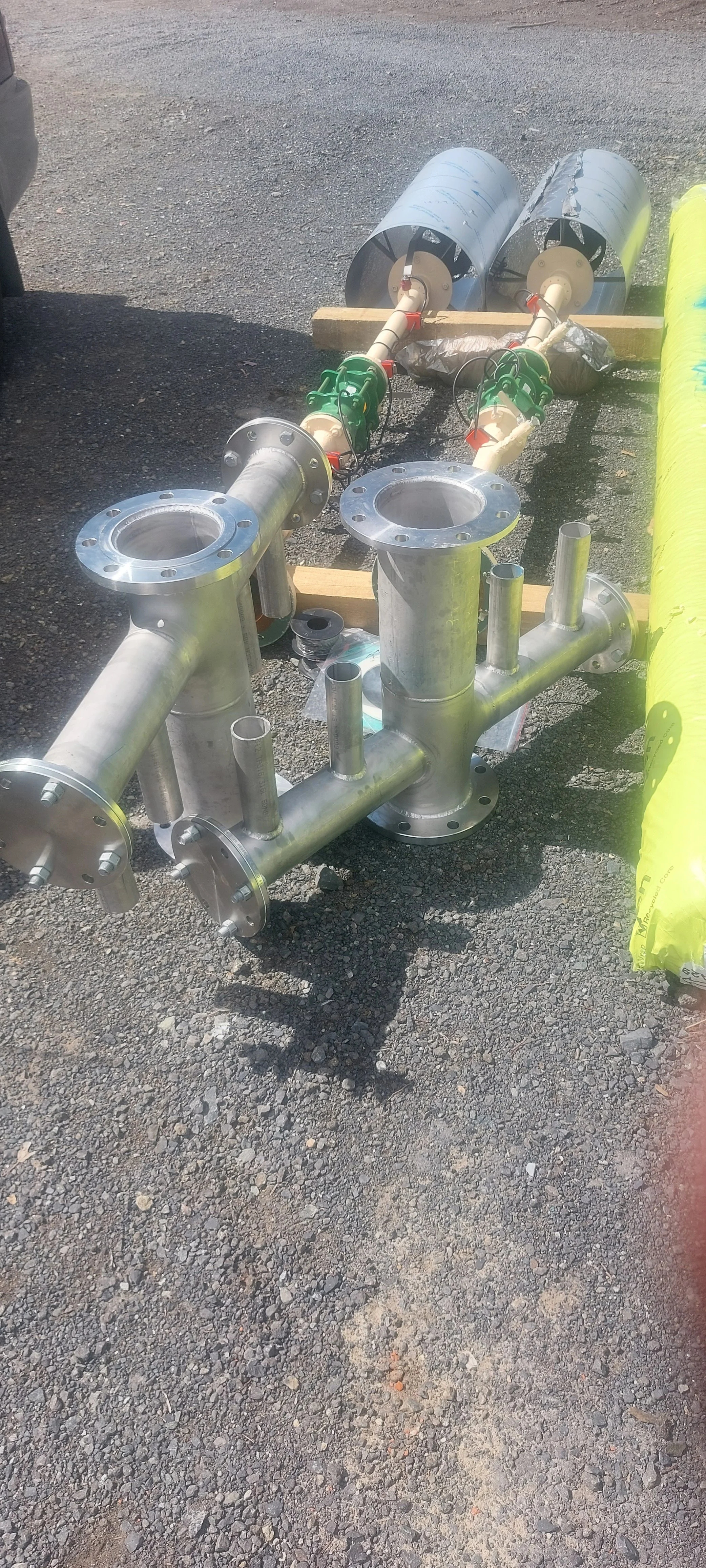 Metal pipeline components, including flanged pipes and valves, lying on gravel ground at an outdoor construction site.