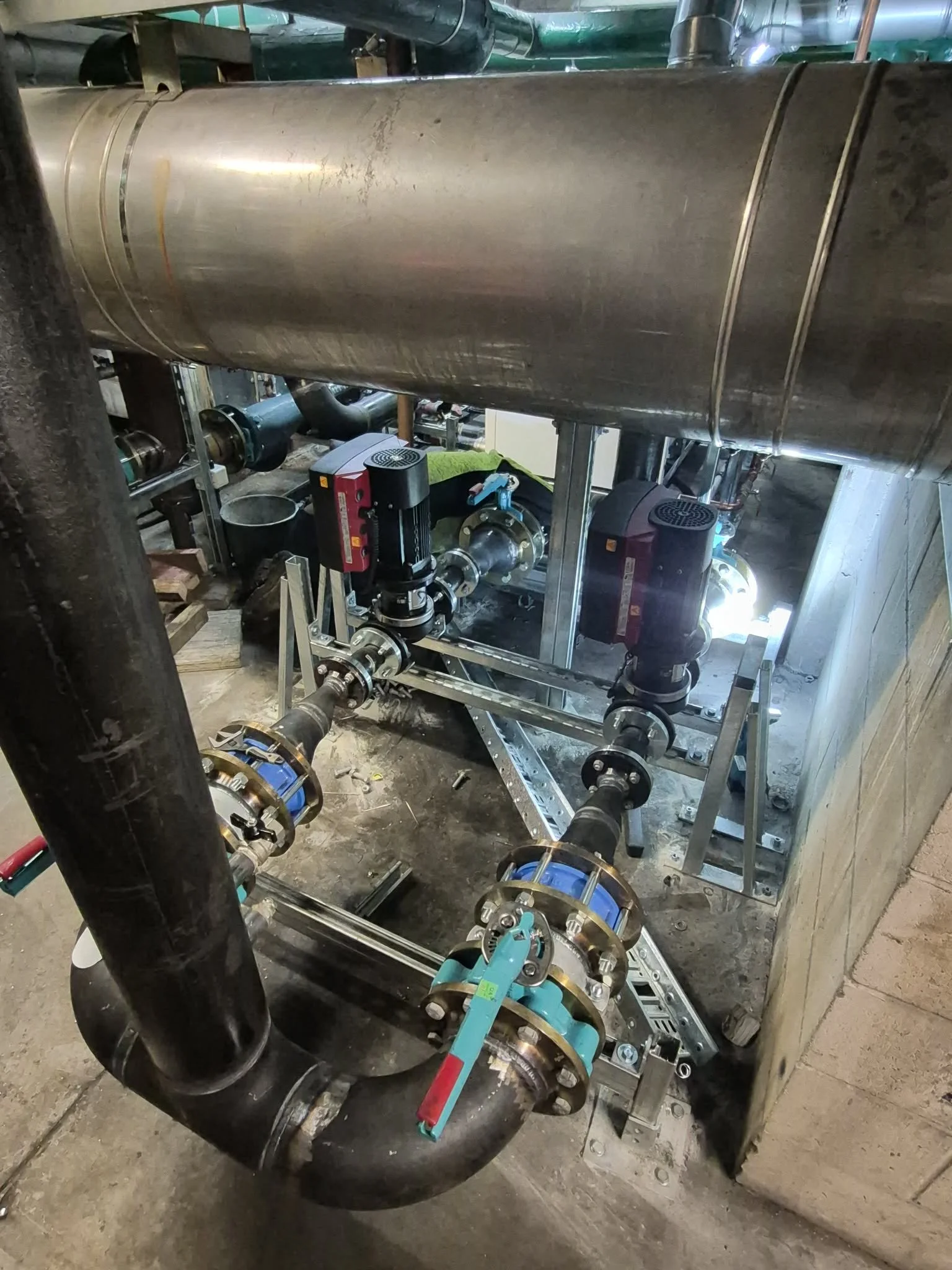 A large metallic cylindrical tank installed within a metal frame with visible pipes, valves, and pumps, likely part of an industrial or mechanical system, in a construction or maintenance setting.