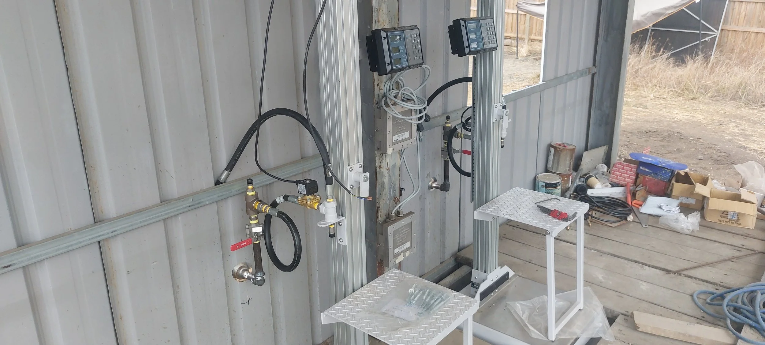 Construction site with tools and equipment, including two control panels, pipes, hoses, a slide hammer, and a work table with tools, set against a metal wall with unfinished wooden flooring.