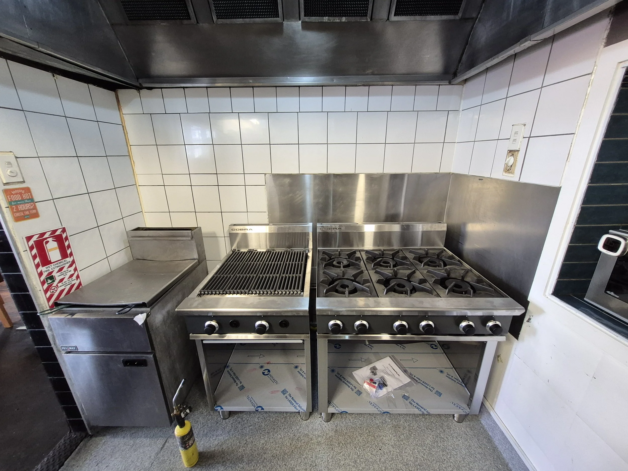 Commercial kitchen with stainless steel appliances including a griddle, a six-burner stove, and a deep fryer, against a tiled wall.