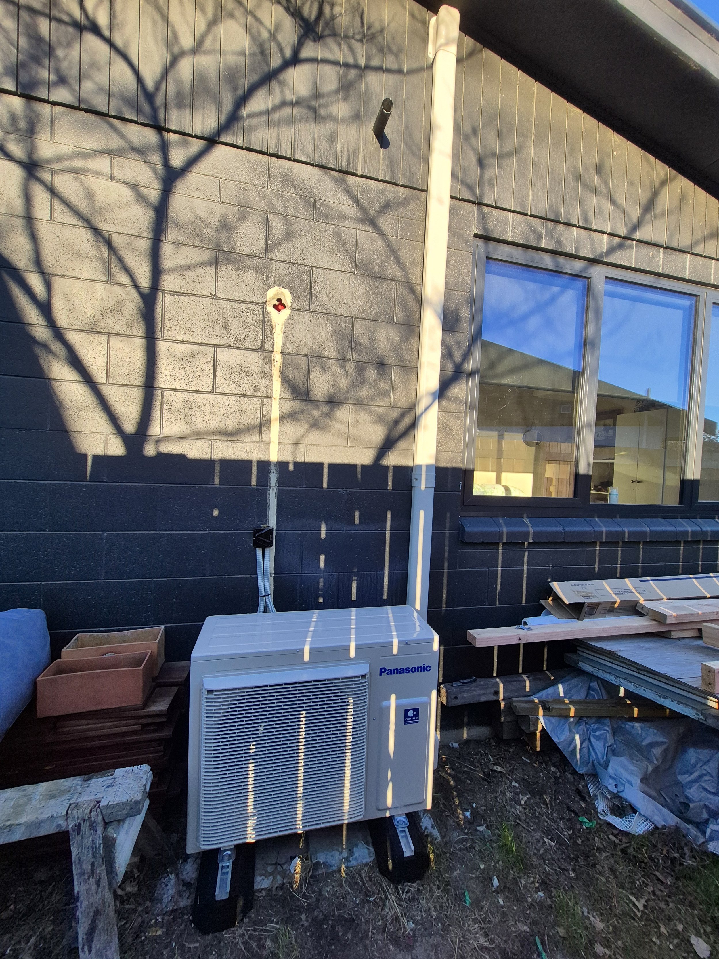 Outdoor scene showing an air conditioning unit, a window with a reflection of the blue sky, a wood bench with a cover, some construction materials, and shadows of tree branches on a gray wall.