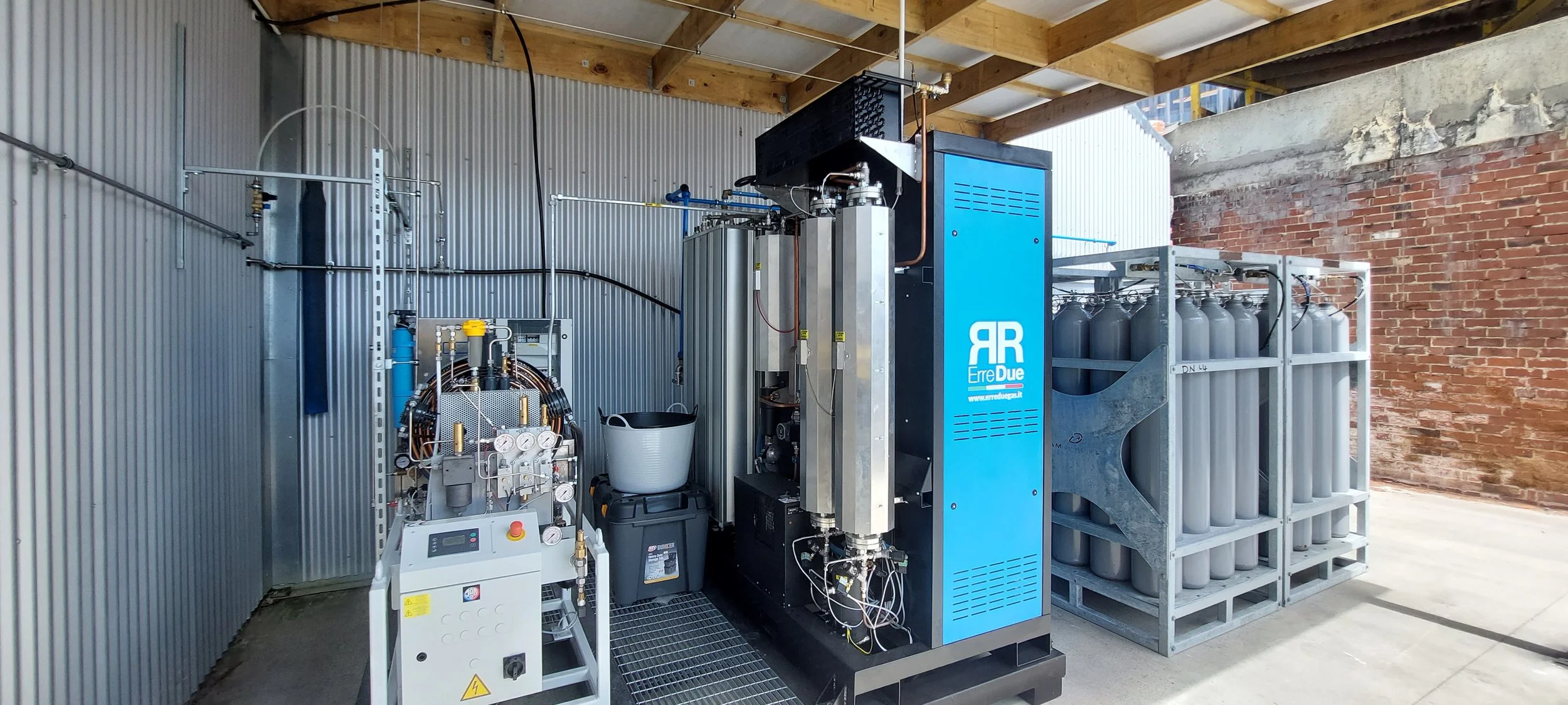 Industrial equipment with gas cylinders on a pallet, various pipes and control panels, located in an open-air covered space with metal walls and a brick wall.