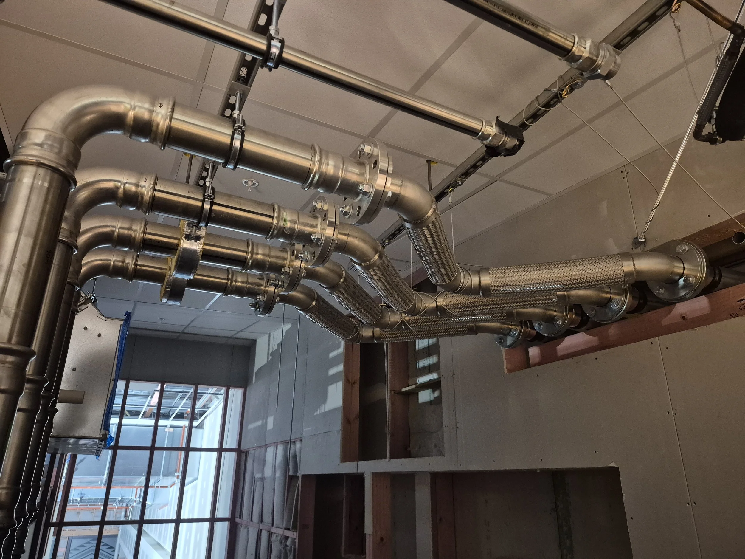 Multiple stainless steel pipes with fittings and insulation running along the ceiling of a building under construction.