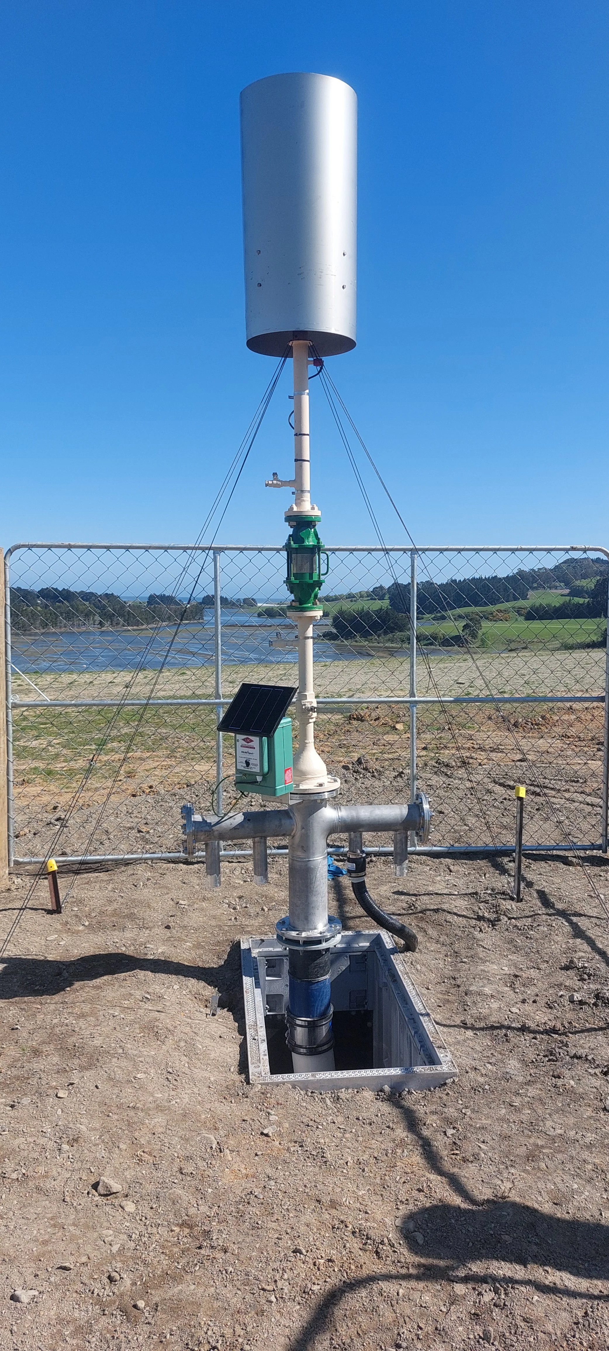 A weather station with a large cylindrical sensor mounted on a pole, surrounded by a fence, with a river and green landscape in the background under a clear blue sky.