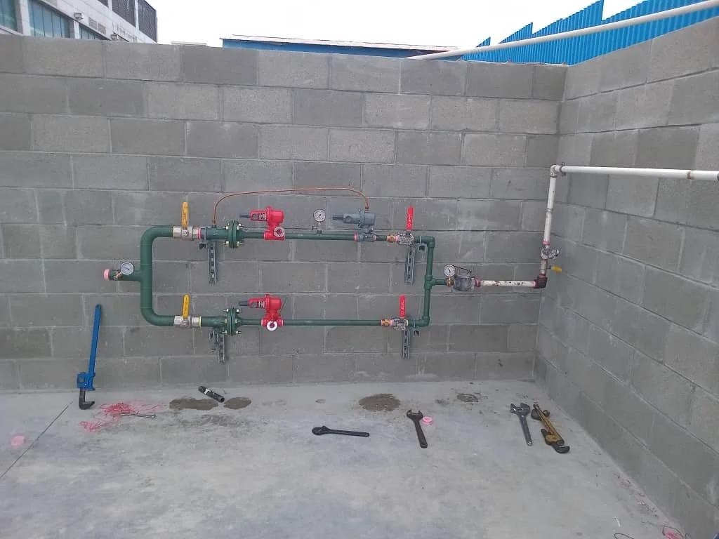The image shows a concrete wall with green and white plumbing pipes, valves, and gauges. Tools are laid on the ground underneath.