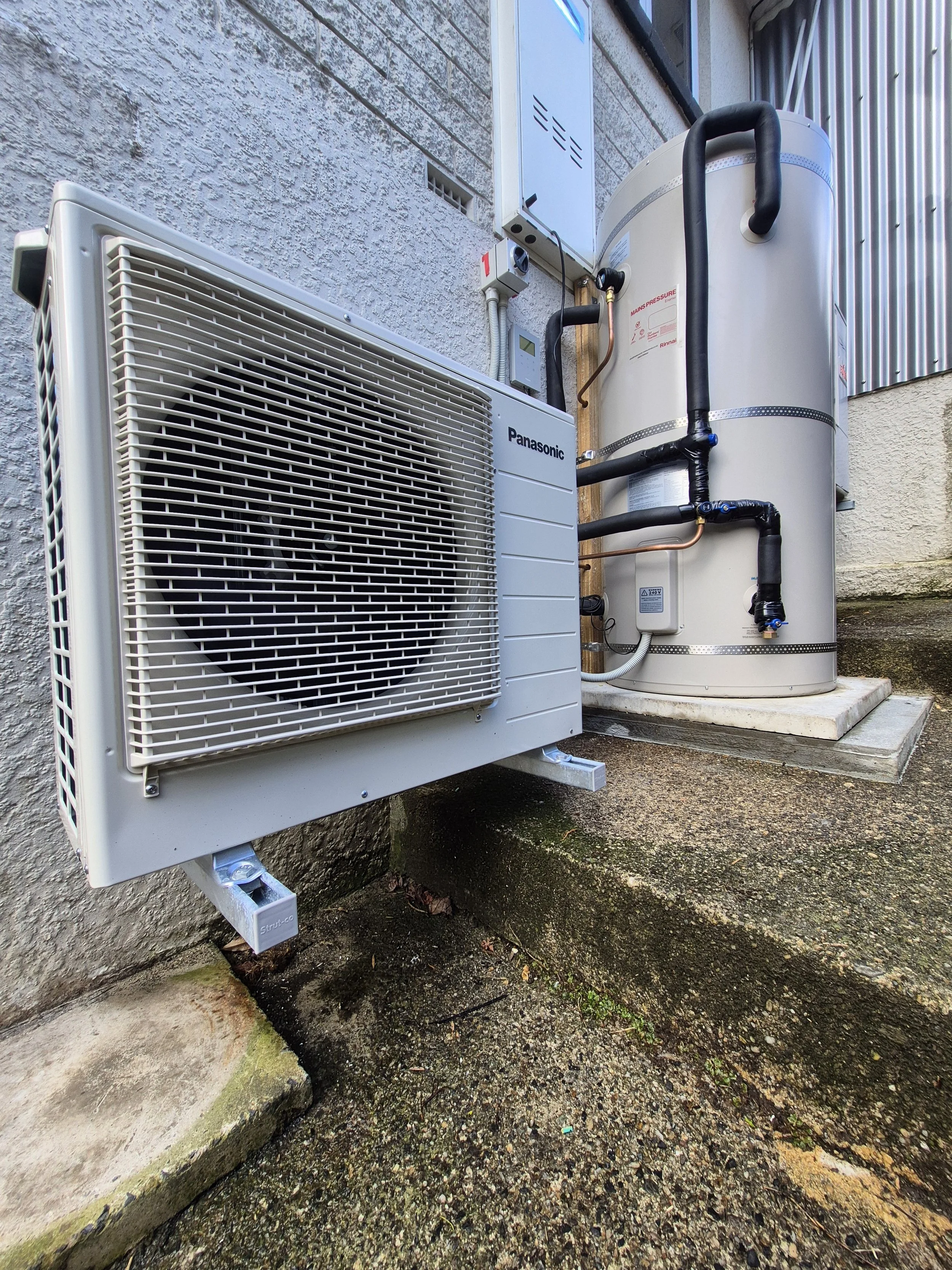 Outdoor view of a wall-mounted air conditioning unit and hot water heater with electrical wiring and pipes. The ground is concrete and some moss and dirt are visible.