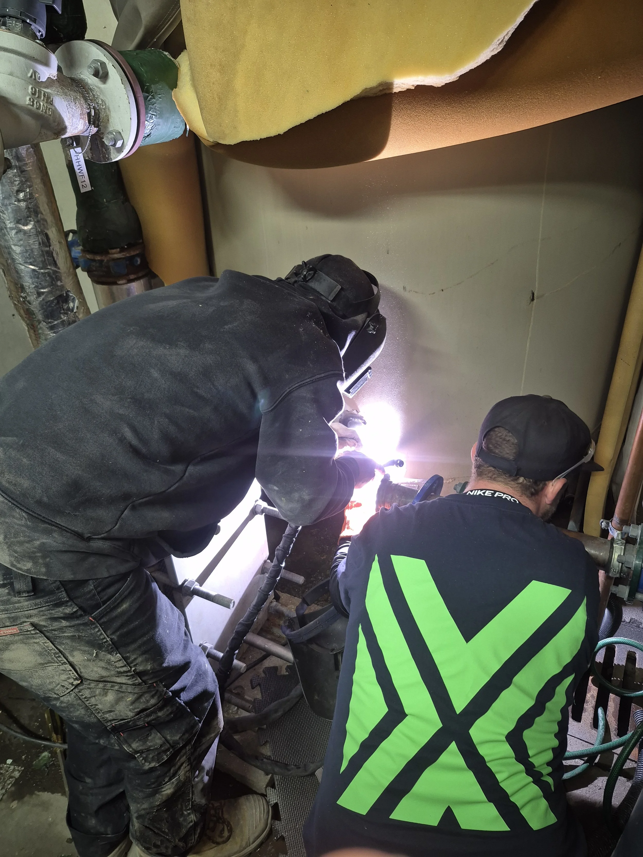 Two workers are welding or repairing a pipe in an industrial setting. One is wearing black pants and a dark shirt with neon green stripes, and the other is wearing a dark hoodie. They are crouching close to the pipe, which is connected to other pipes