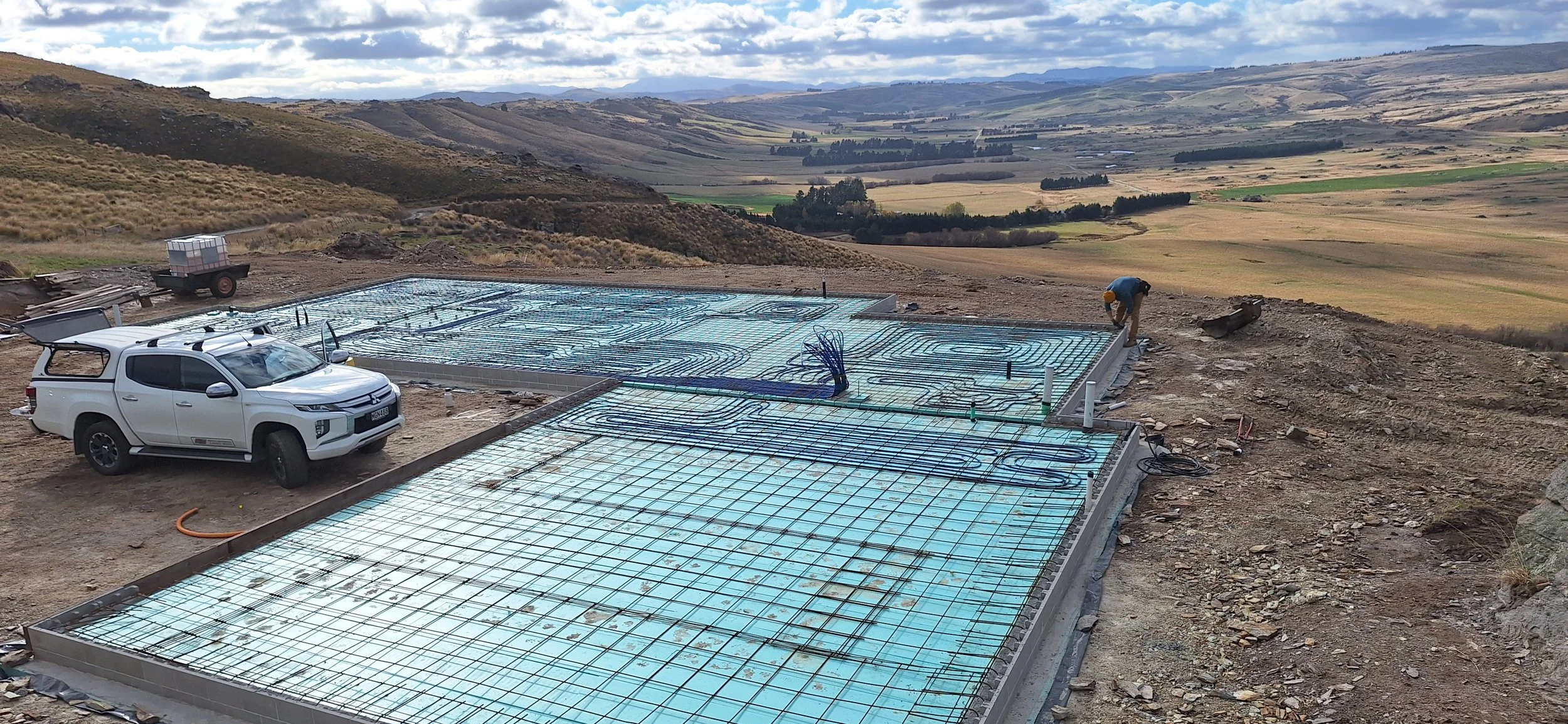 Construction site in a rural landscape with a foundation being prepared for building, including exposed rebar and a worker installing plumbing or electrical lines, and a vehicle parked nearby under a cloudy sky.