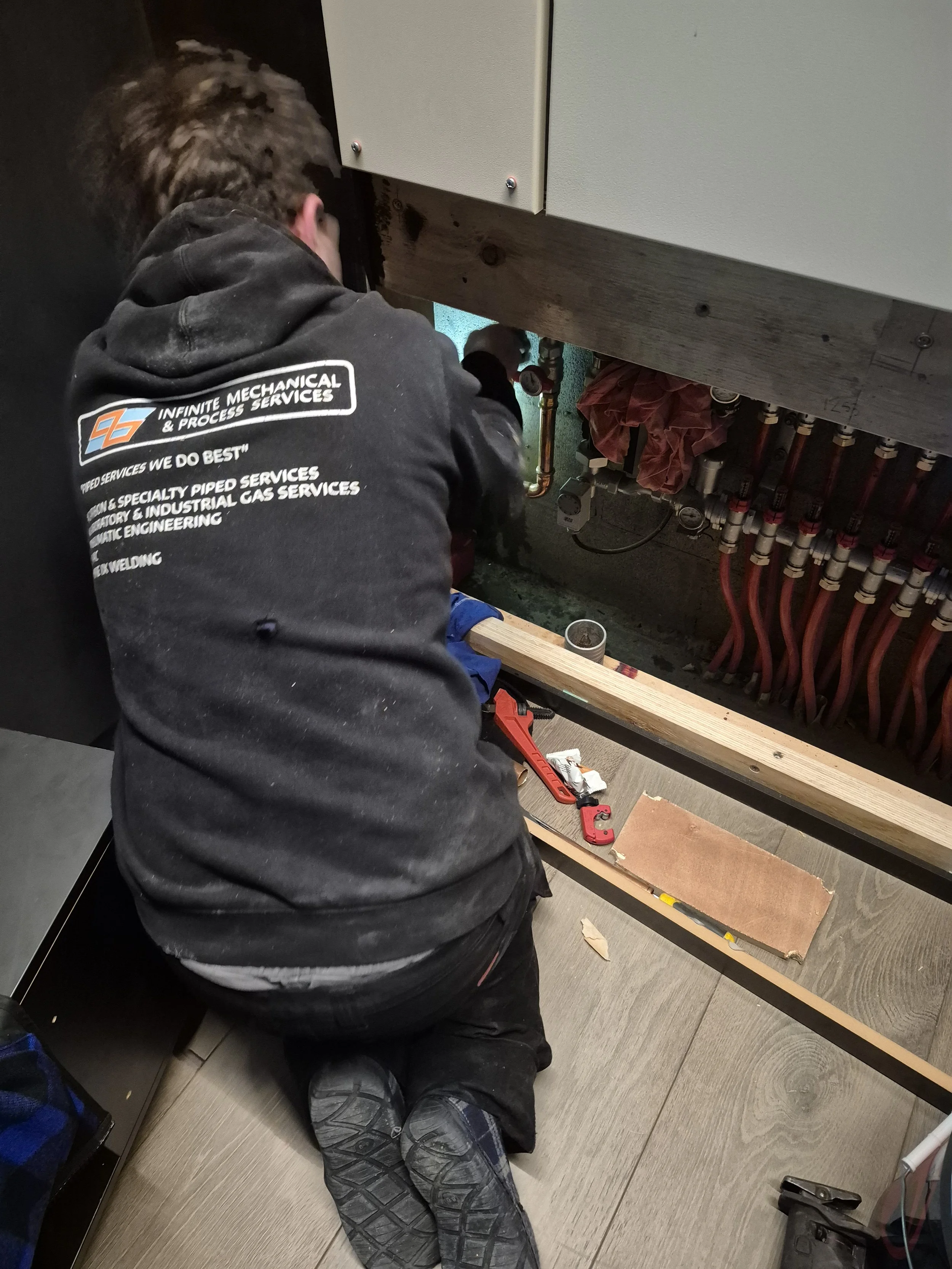 A person kneeling on the floor working on plumbing under a sink, wearing a black hoodie with the logo 'Infinite Mechanical & Process Services' on the back and black work boots.