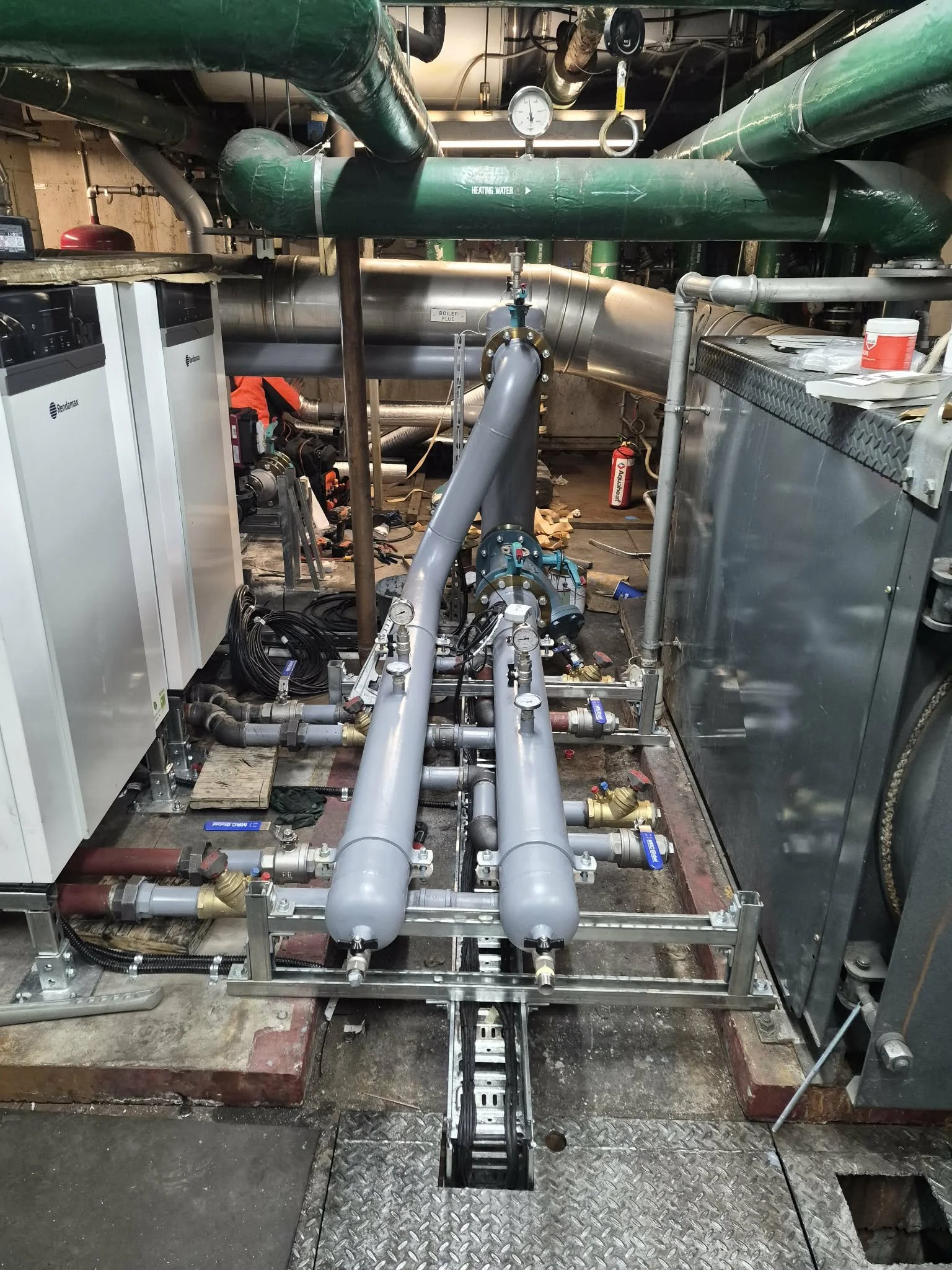 View of industrial mechanical equipment and piping, including large green pipes labeled 'Heating Water,' metal valves, and gauges, within a utility or mechanical room.