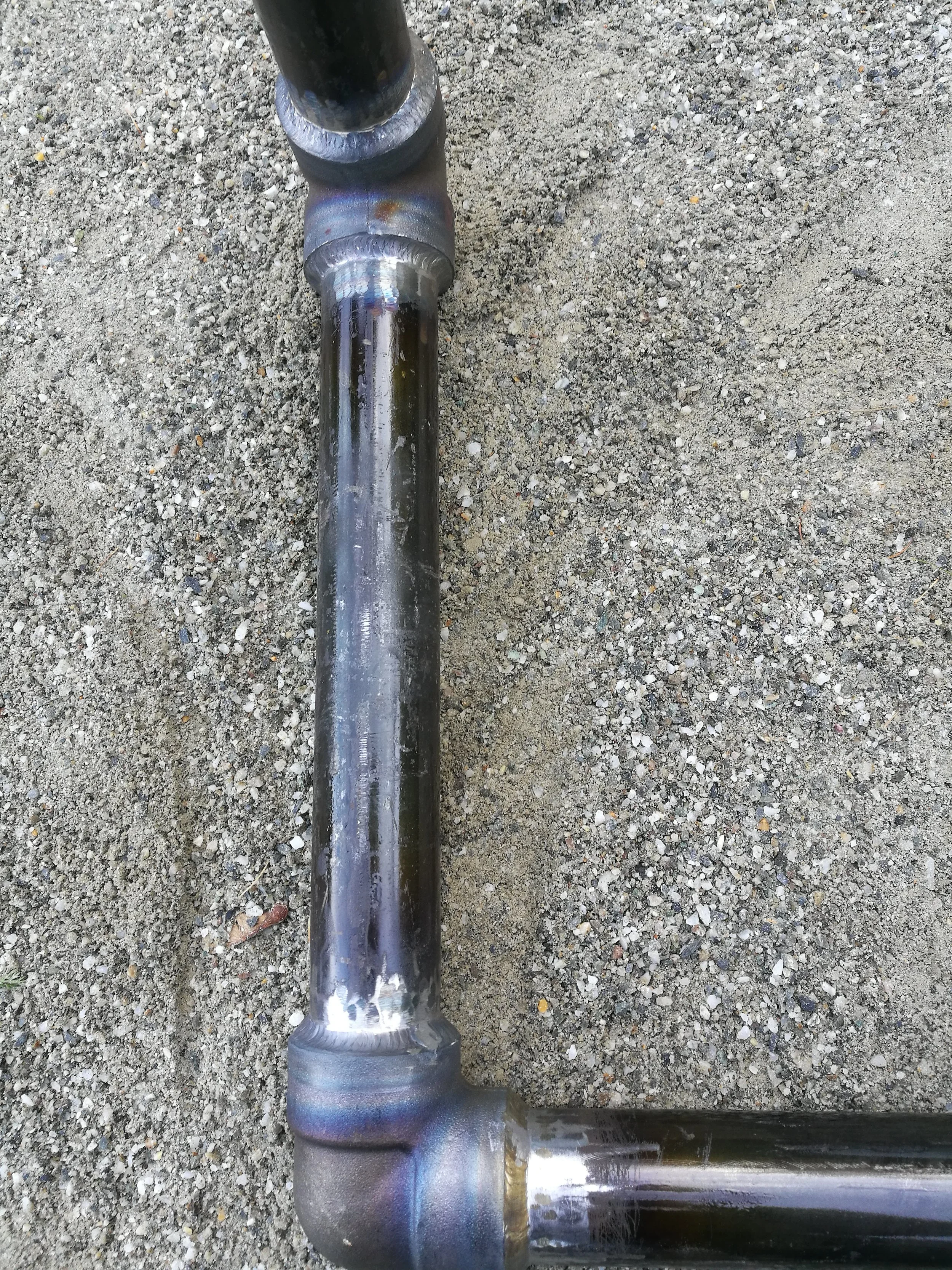Close-up of welded metal pipes on a gravel surface, showing weld beads and discoloration from welding heat.
