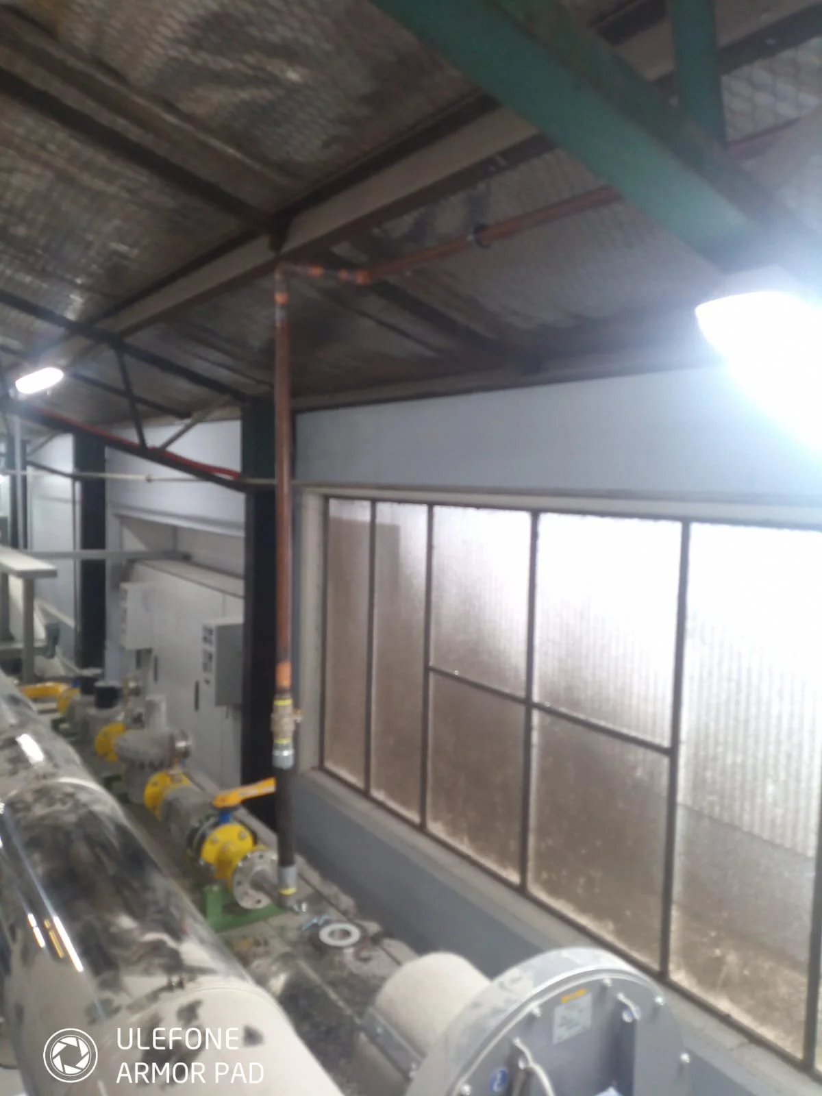 Industrial interior with metal pipes and machinery, frosted glass windows, and fluorescent lighting.
