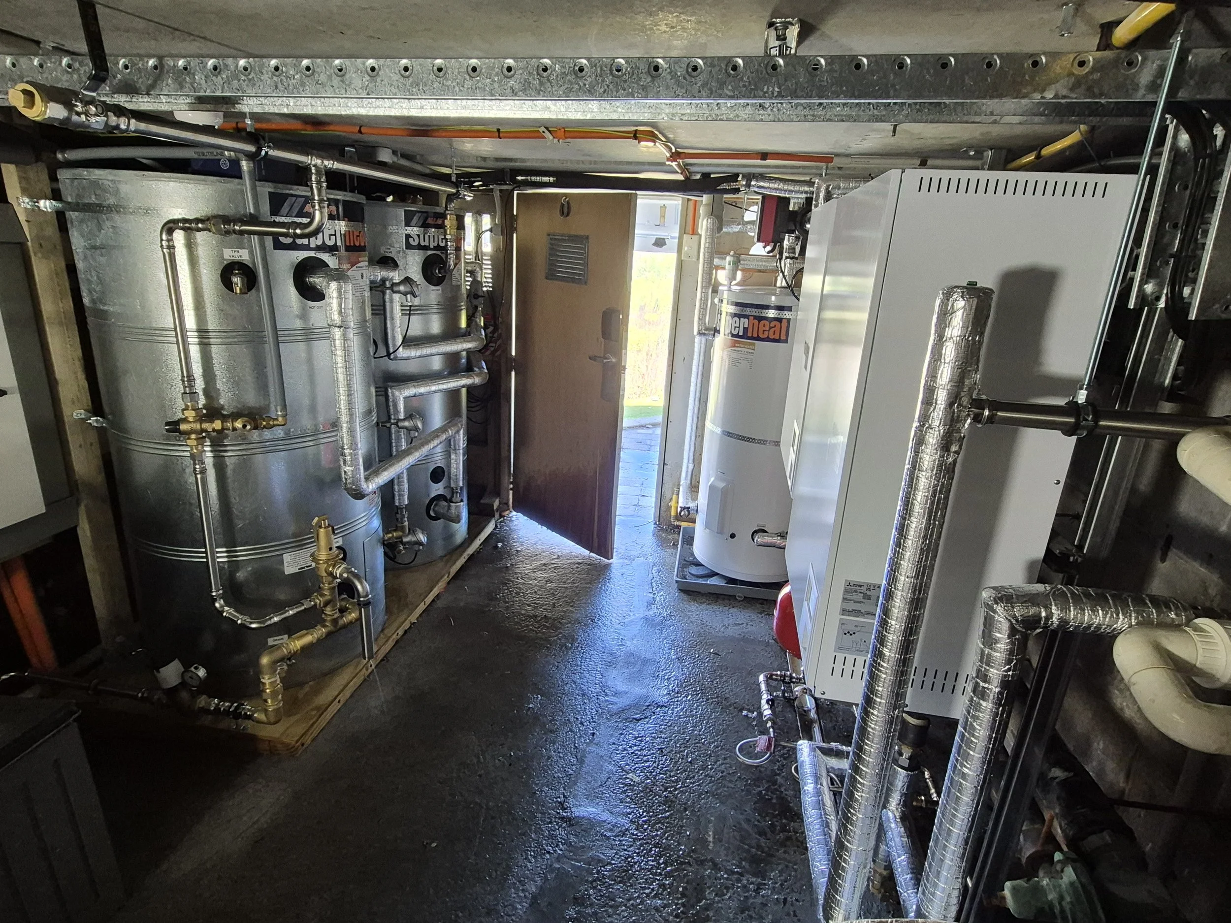 An indoor area with heating and ventilation equipment, including a large water heater, metal pipes, and insulation, with a partially open door leading outside.