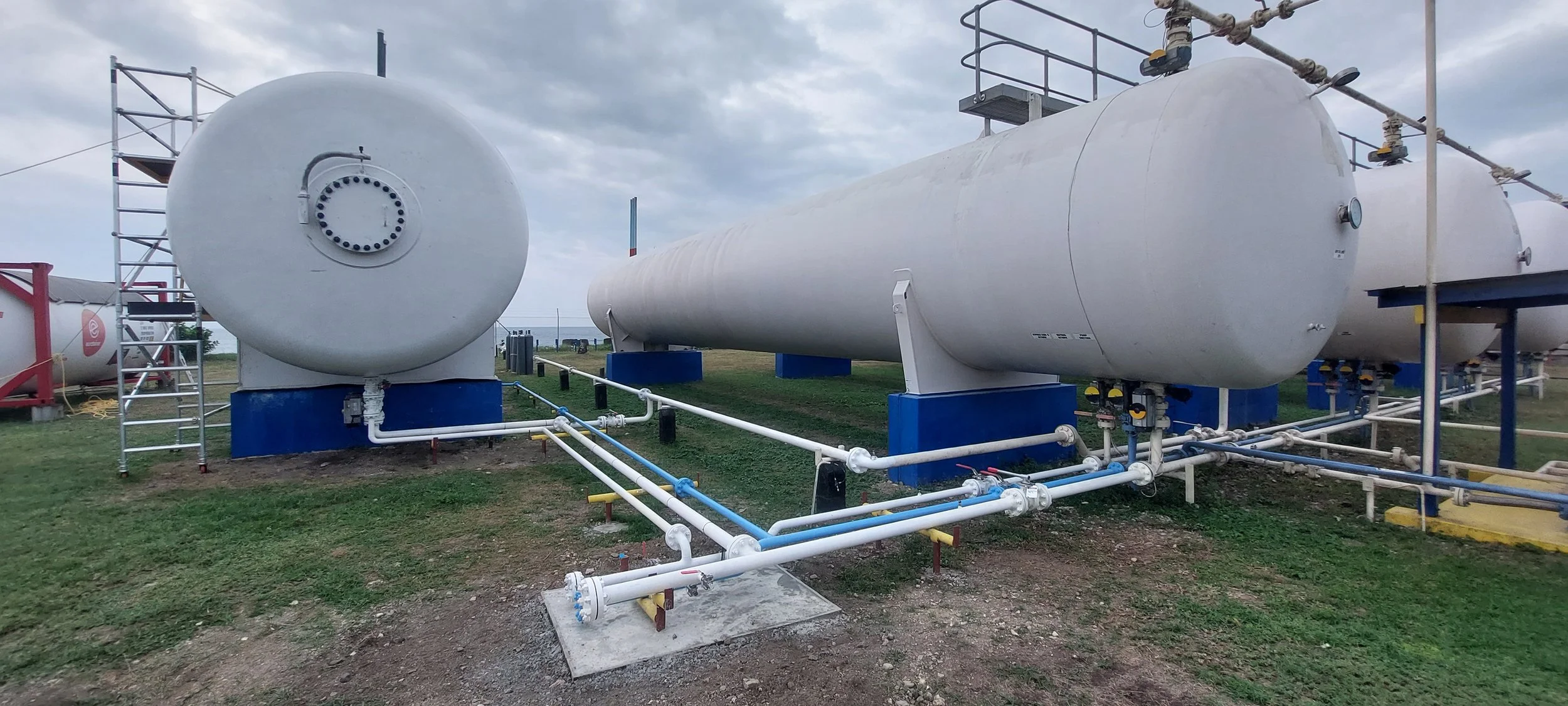 Large white cryogenic storage tanks on a grassy field with pipelines and a metal staircase, under a cloudy sky.