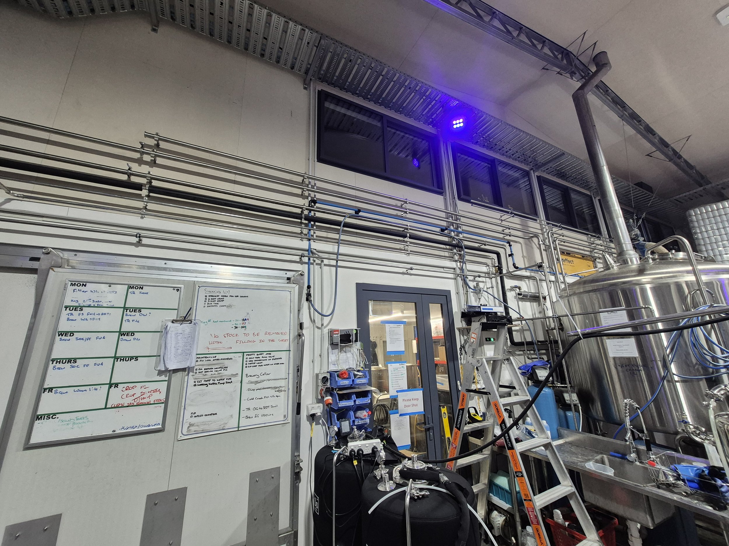 The image shows an industrial or laboratory setting with stainless steel equipment, pipes, and tanks. There are also ladders, tools, and a whiteboard with handwritten notes.