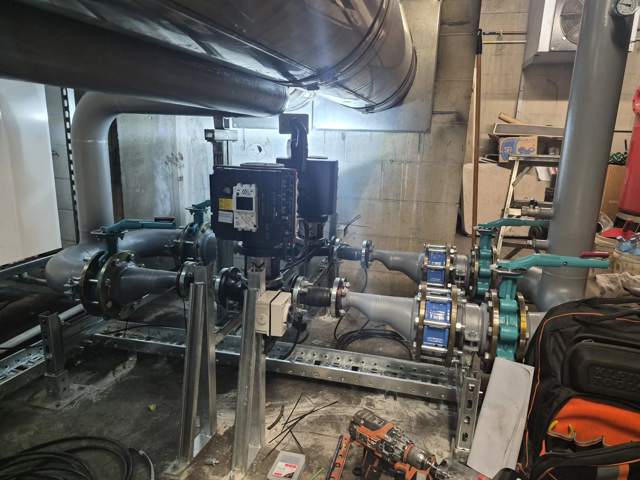 Underground utility or mechanical room with pipes, valves, and equipment, including a hand-held drill, toolbox, and miscellaneous tools.