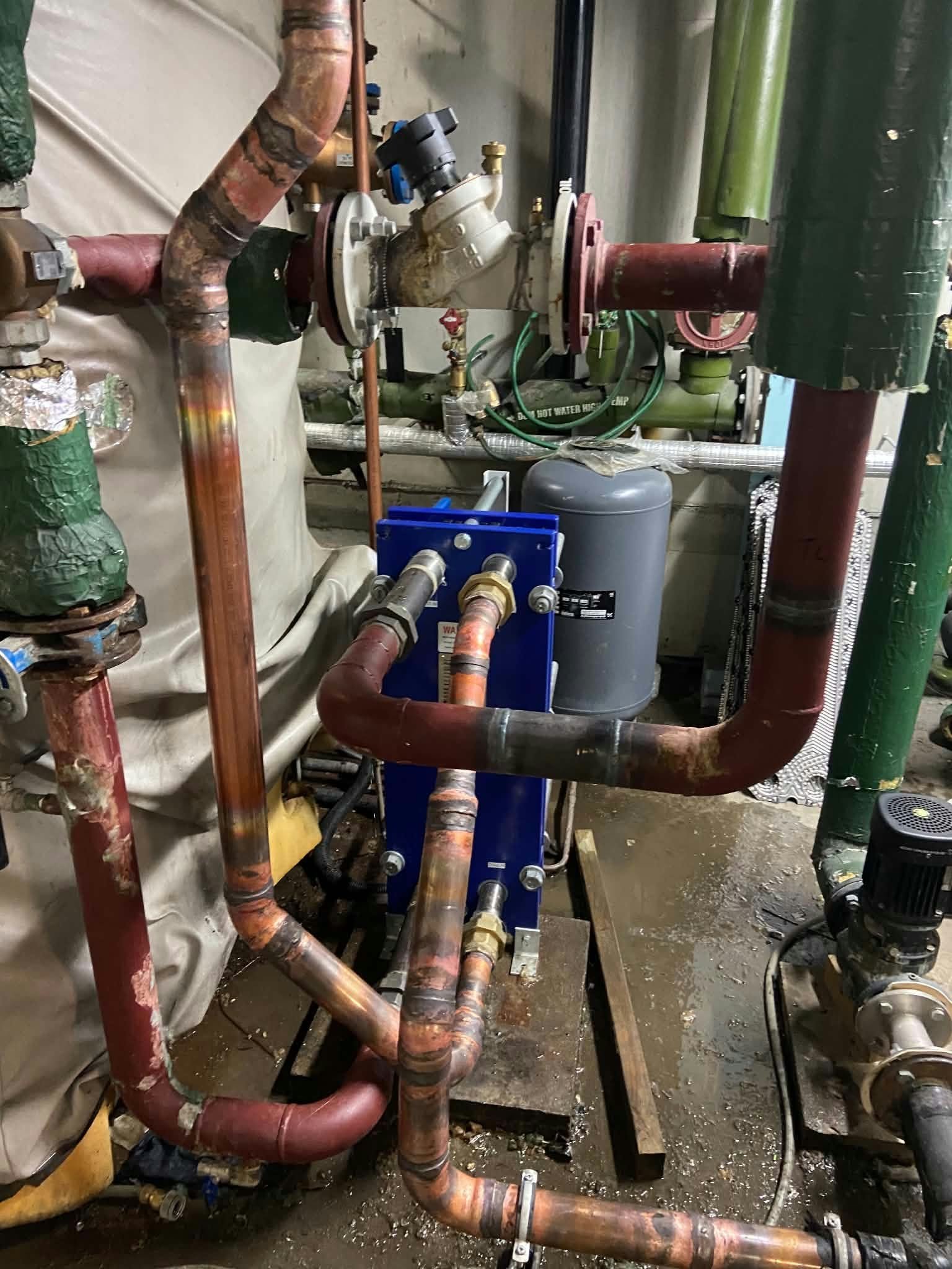Photo of plumbing pipes and equipment in a mechanical room, some pipes are red and copper, with valves and fittings, and a gray electrical box is visible.