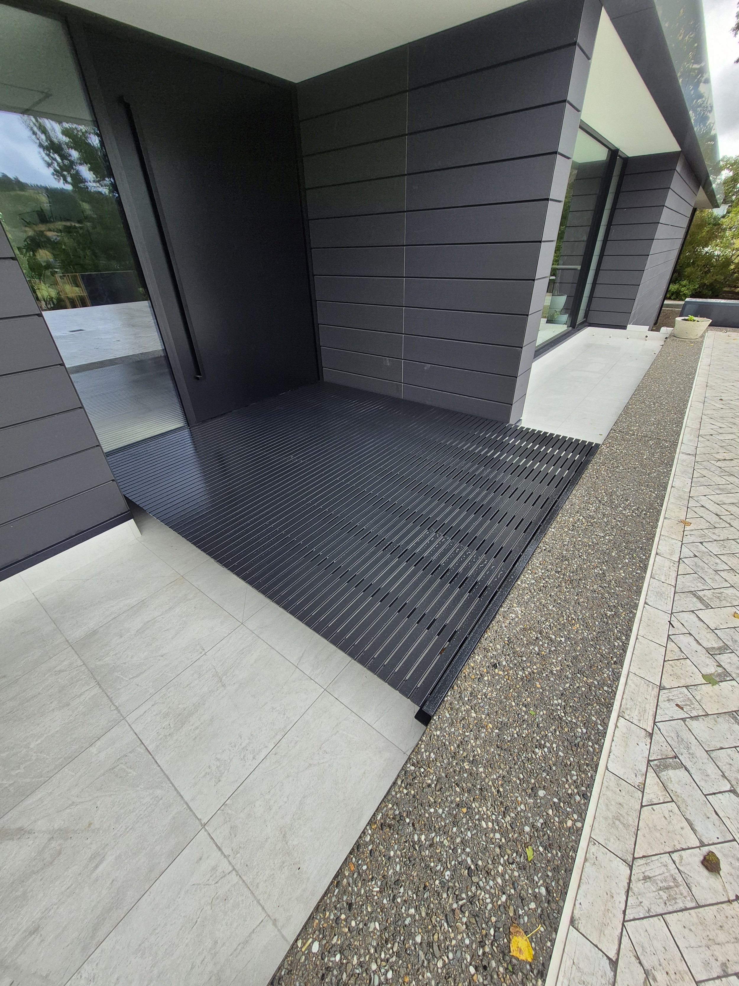 Modern house entrance with black metal grate walkway, light gray stone tiles, and black siding.