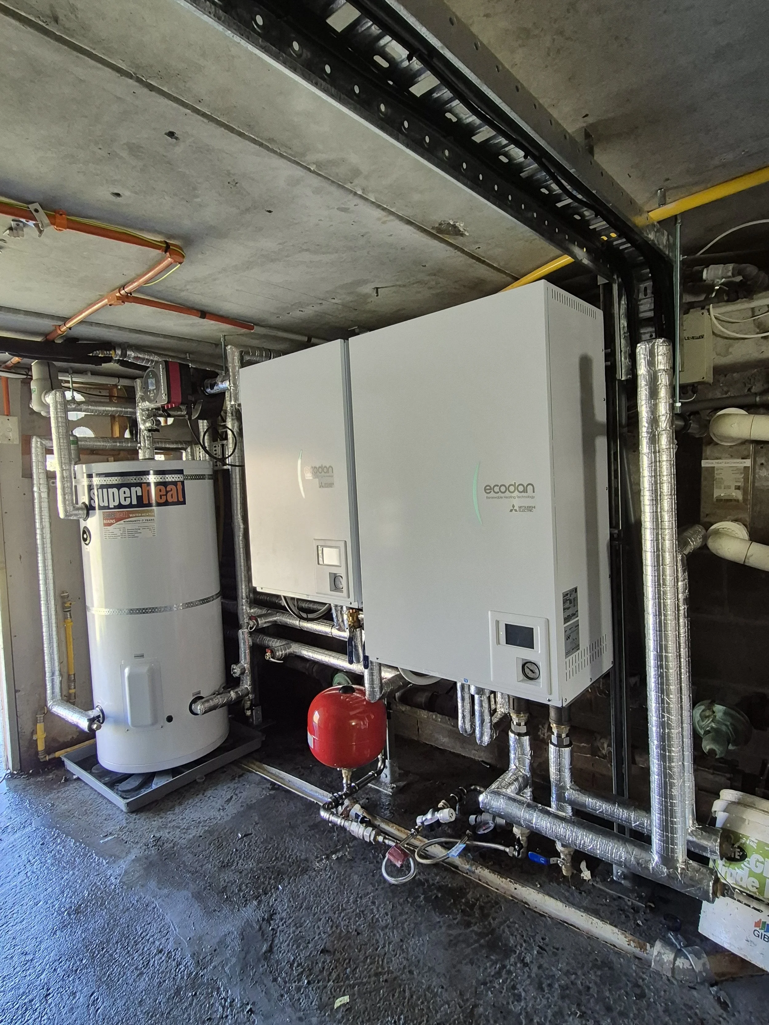 A utility room with a water heater and a large white HVAC or boiler system wrapped in insulation, with various pipes, valves, and electrical components connected to it. The floor is concrete, and there are pipes along the wall and ceiling.