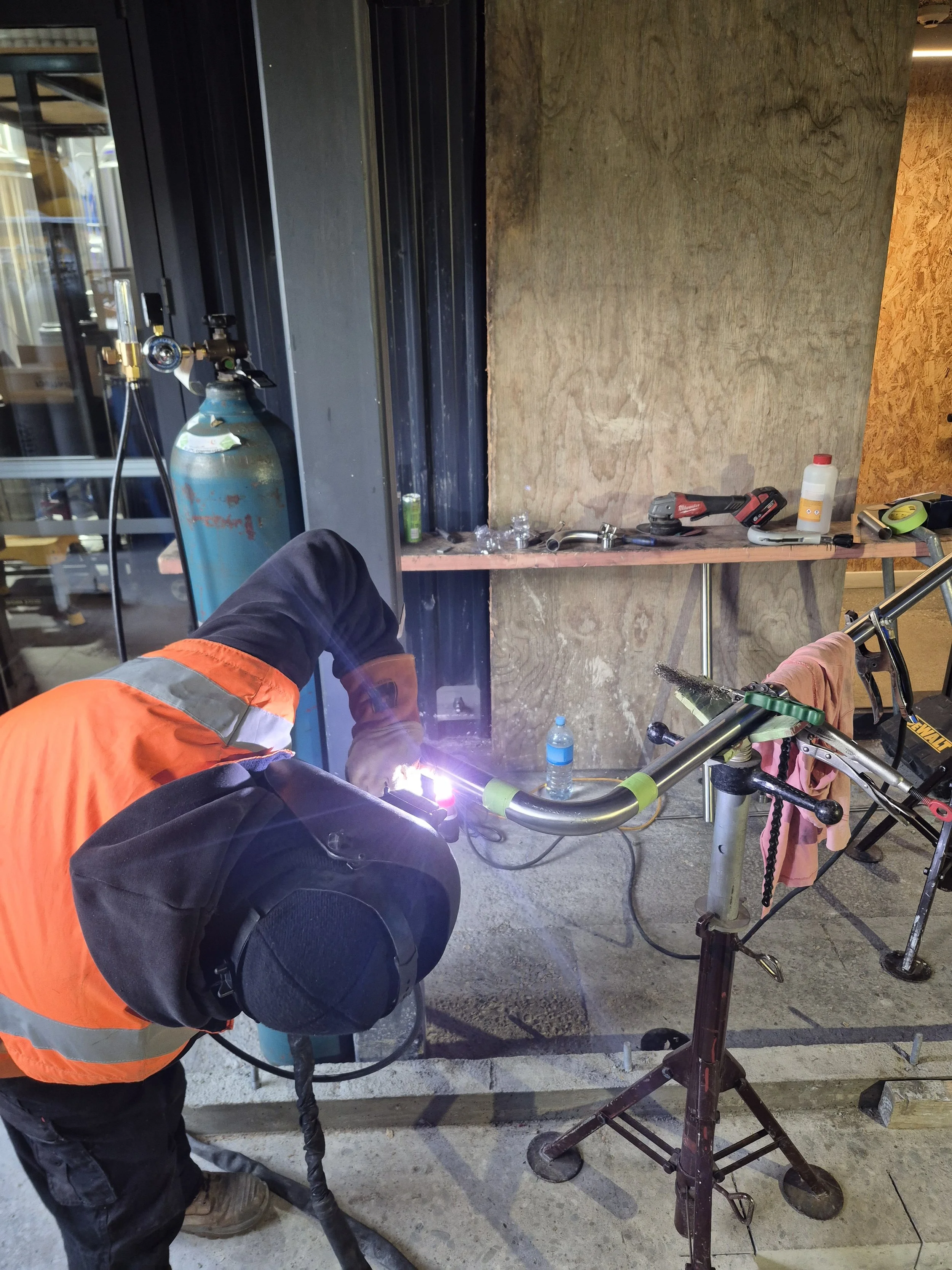 A worker in an orange safety vest welding metal pipes and parts in an industrial or construction setting. The worker is wearing gloves and is focused on the welding process. There are tools, water bottles, and construction materials visible on a wood