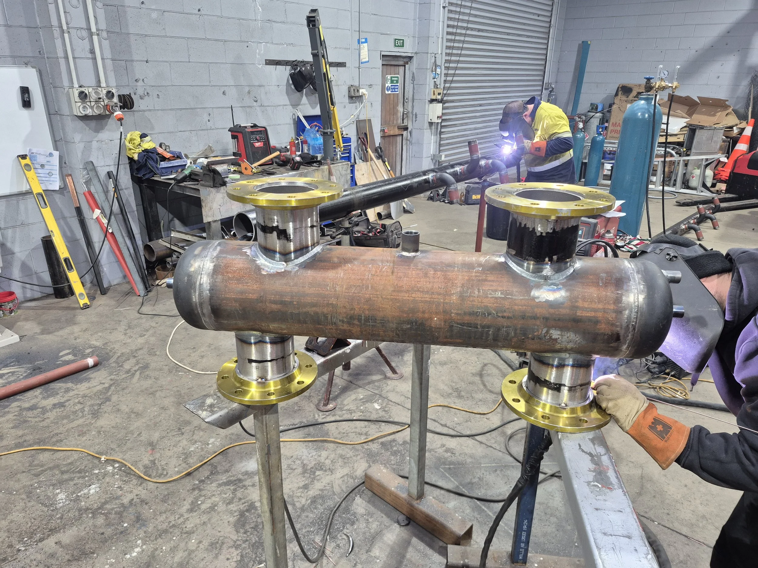 A worker welding a metal pipe in an industrial workshop with tools and equipment around.