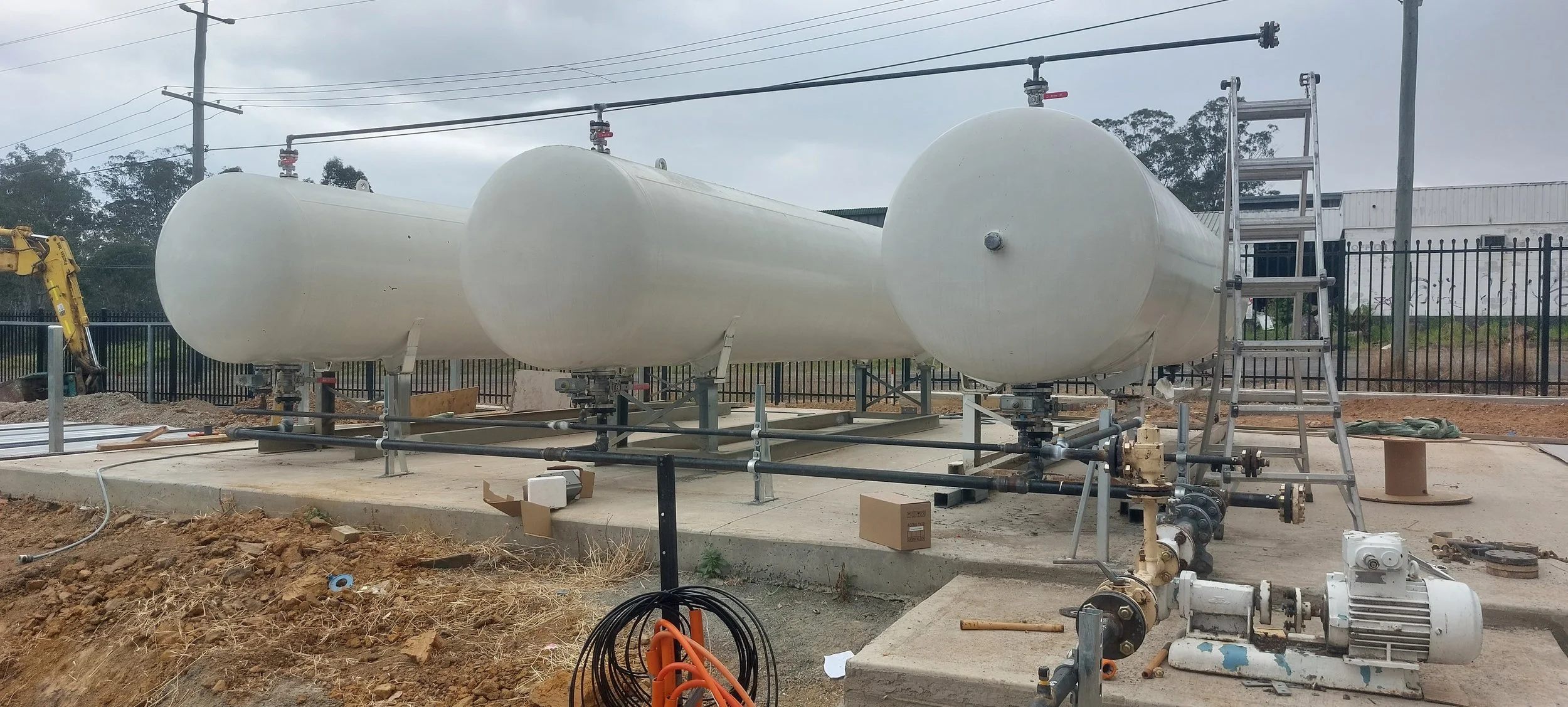 Three large white underground storage tanks on a construction site, connected by pipes, with a ladder nearby and construction equipment around.
