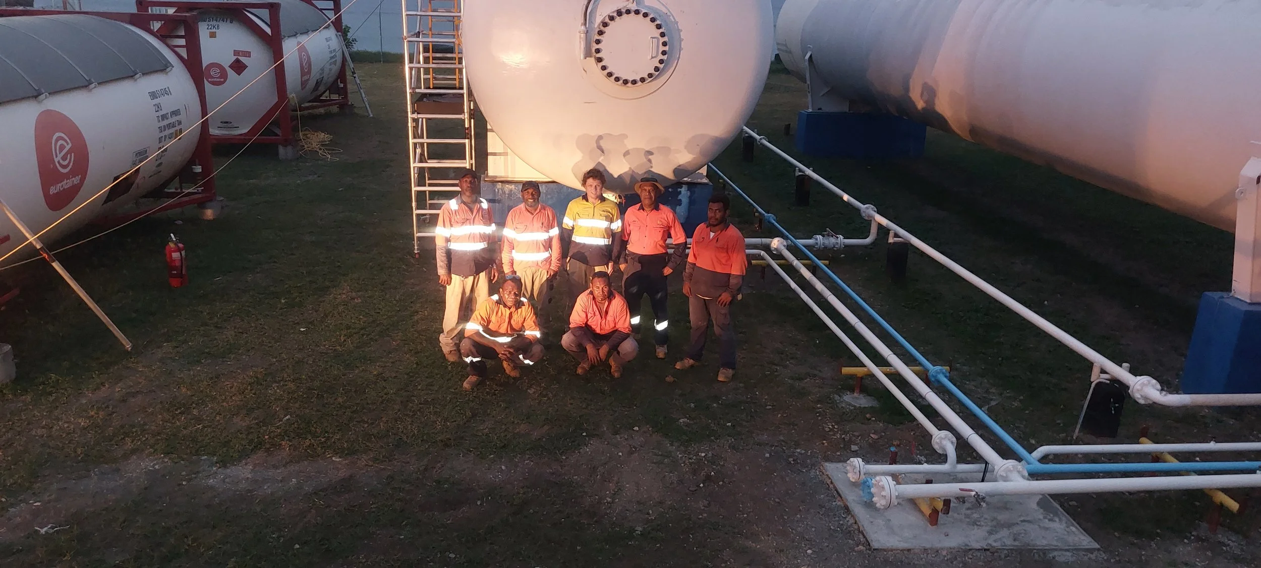 A group of eight workers standing and kneeling in front of large industrial tanks and pipes outdoors during dusk.