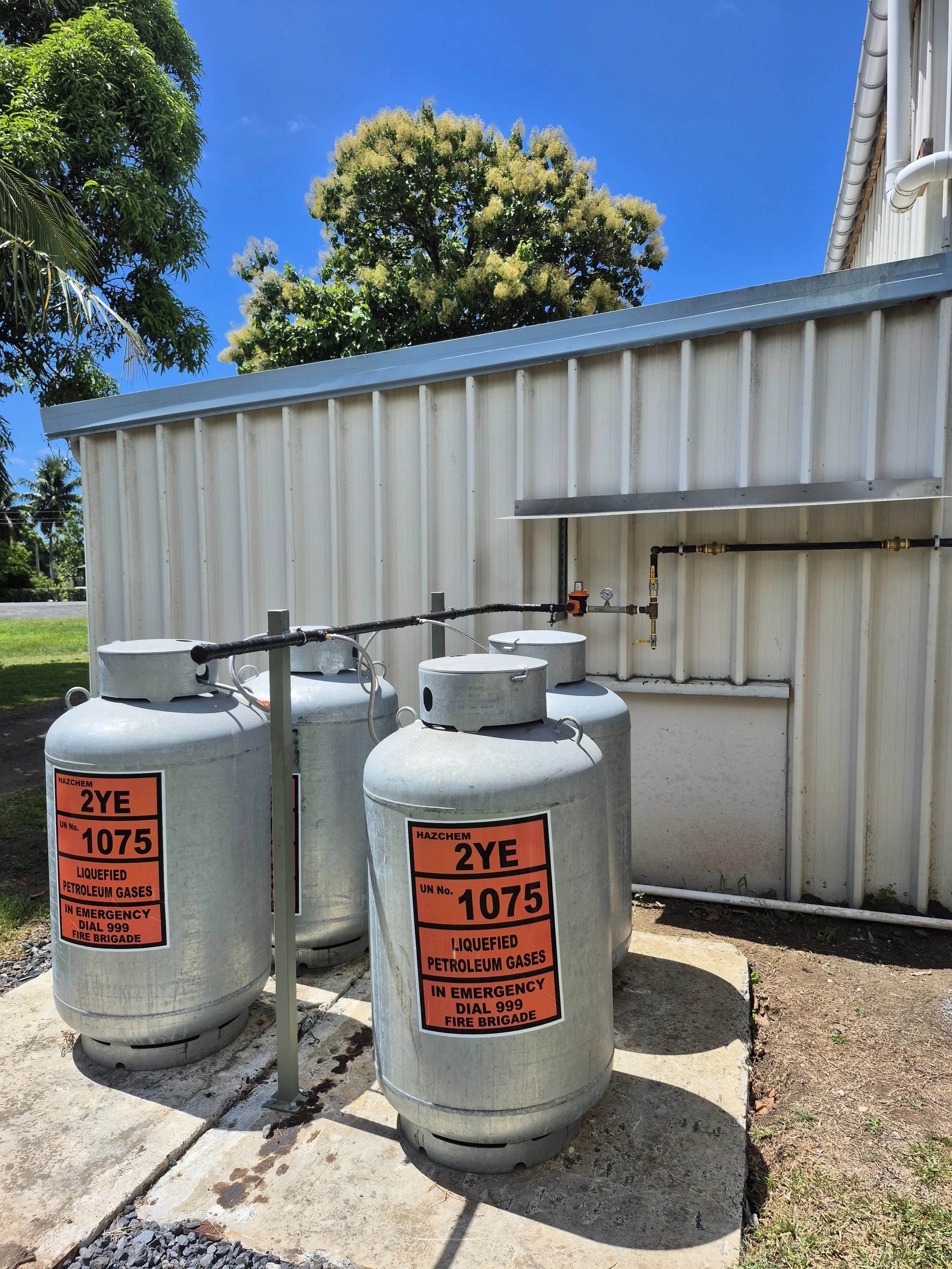 Three large cylinders labeled as liquefied petroleum gases in an outdoor area, with a metal wall and green trees in the background under a clear blue sky.