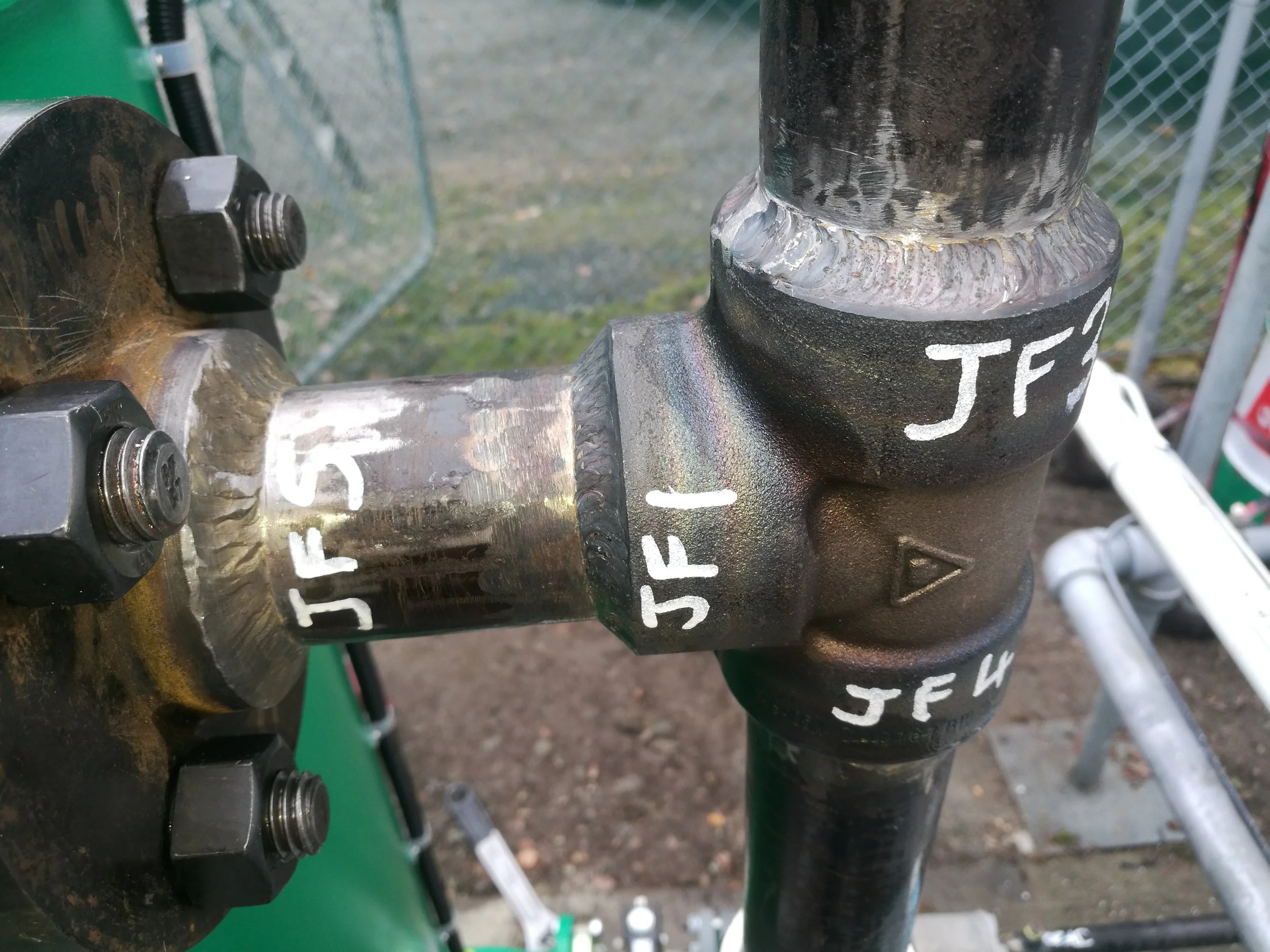 Close-up of welded metal components with visible bolts and a black painted surface marked with white handwritten labels. The background shows a chain-link fence and outdoor setting.