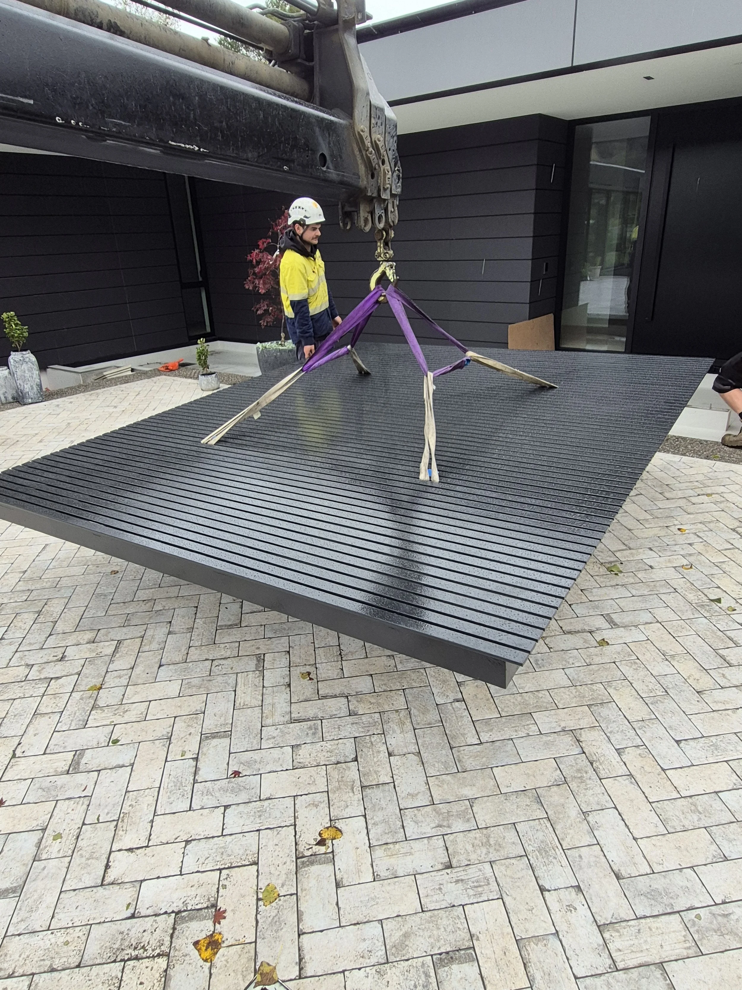 A person in a yellow safety jacket and helmet is operating a crane to lift a large, black, slatted platform, which is suspended in the air in front of a building with dark exterior walls.