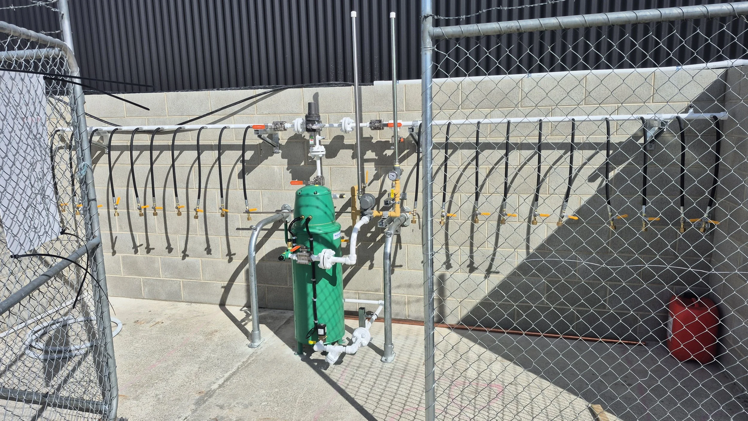 Outdoor water connection system with pipes, valves, and a green pressure tank, enclosed by chain-link fence with a black building in the background and a red container in the corner.