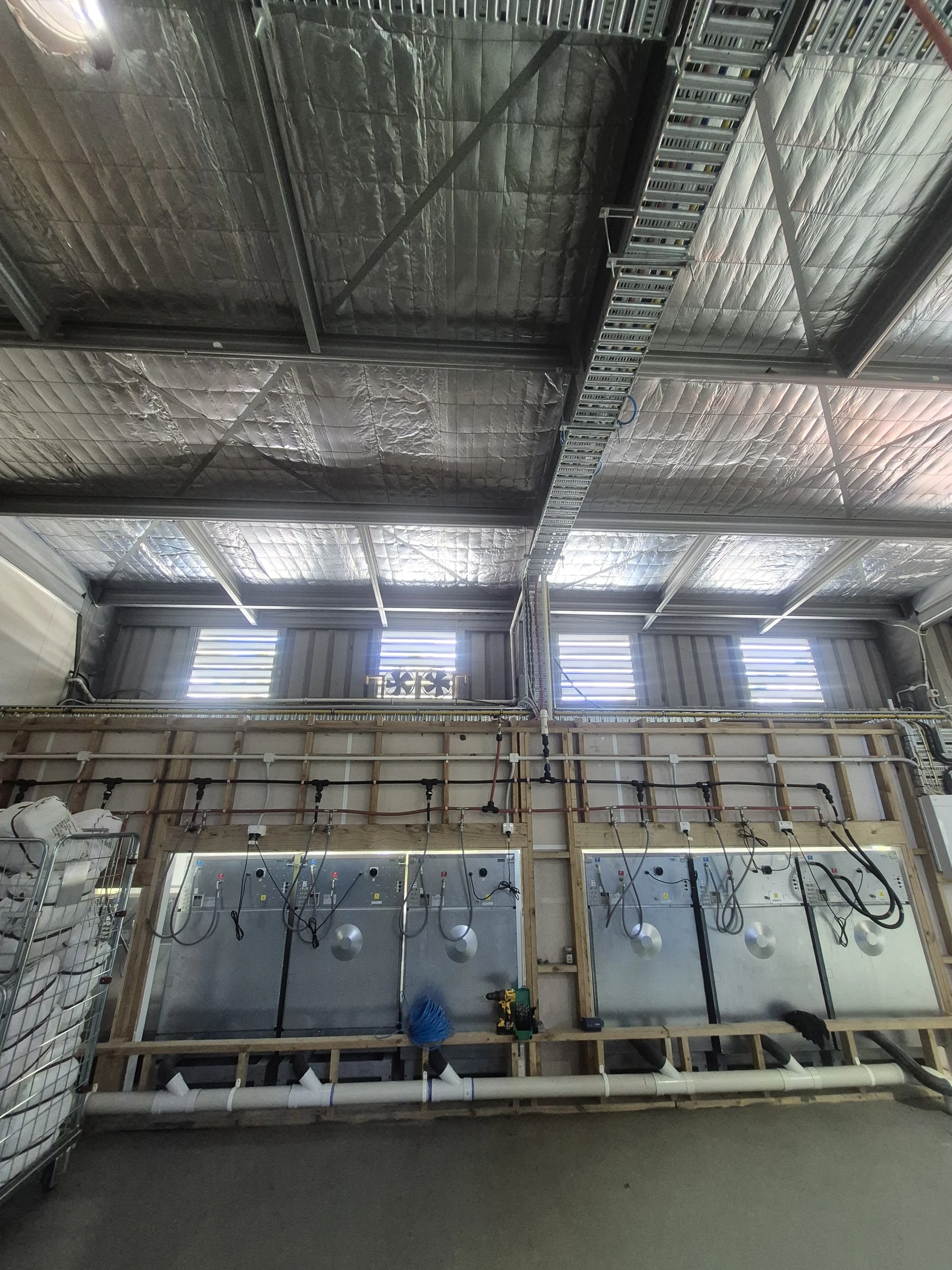 Industrial building interior with exposed metal roofing, insulation, and visible wiring and piping on a wall under construction.