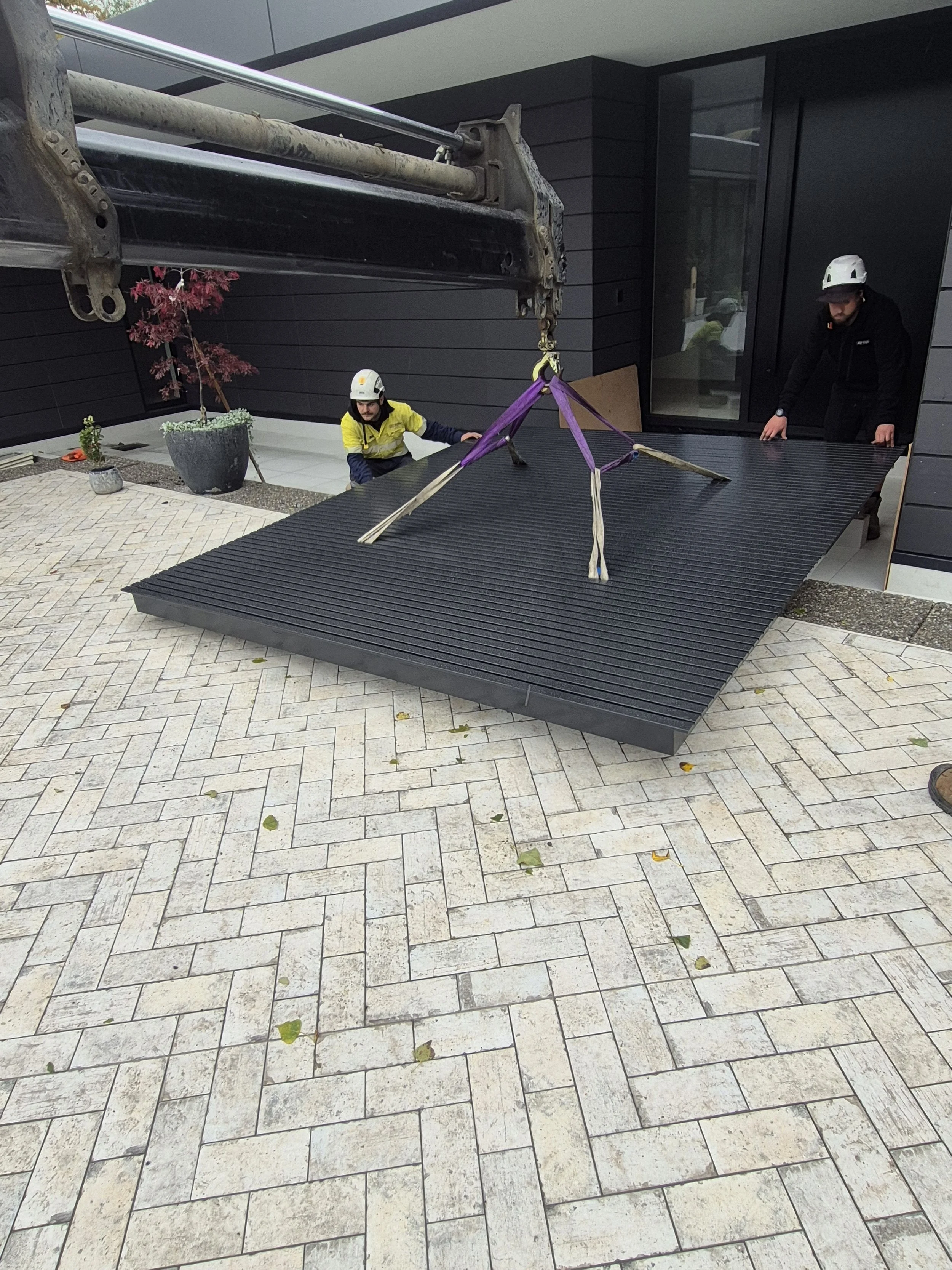 Two construction workers with helmets are installing a large, black, textured platform outside a modern building with dark gray walls. One worker is holding and positioning the platform while the other observes. A crane arm is lifting the platform wi