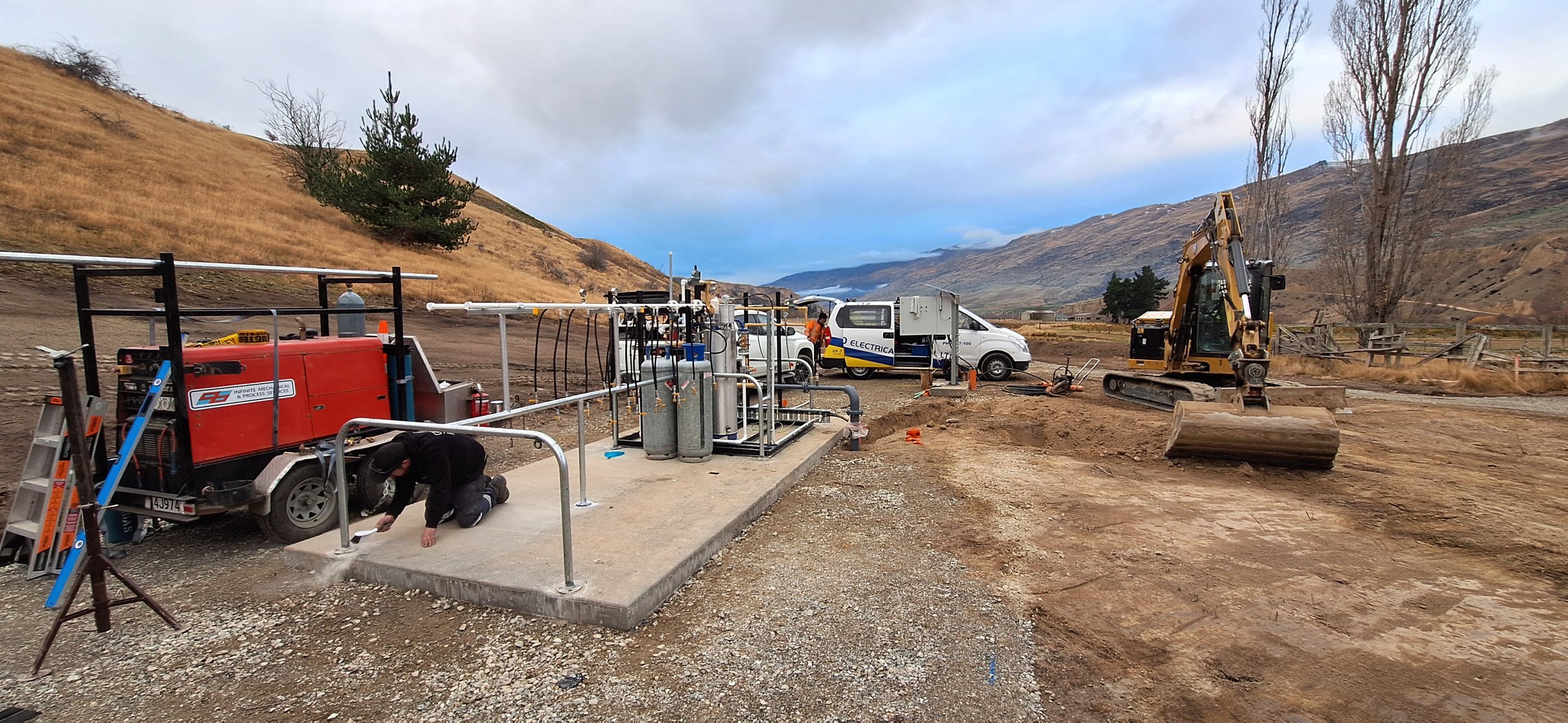 Construction site in a hilly, rural area with equipment, vehicles, and a worker kneeling near machinery, and a small excavator on the dirt ground.